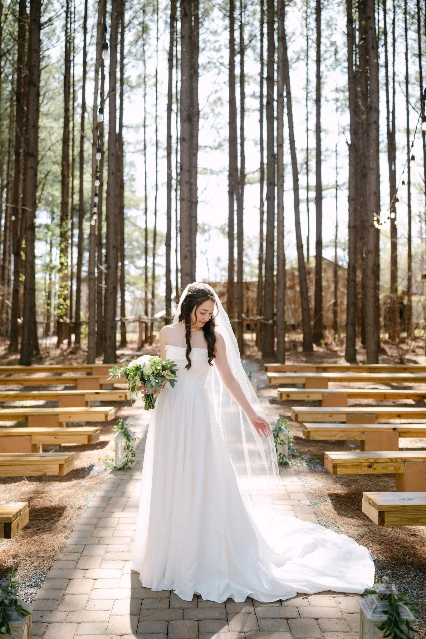Bride holding a white and green bouquet on the brick ceremony aisle beneath tall pine trees at Bishop Farm Event Farm.