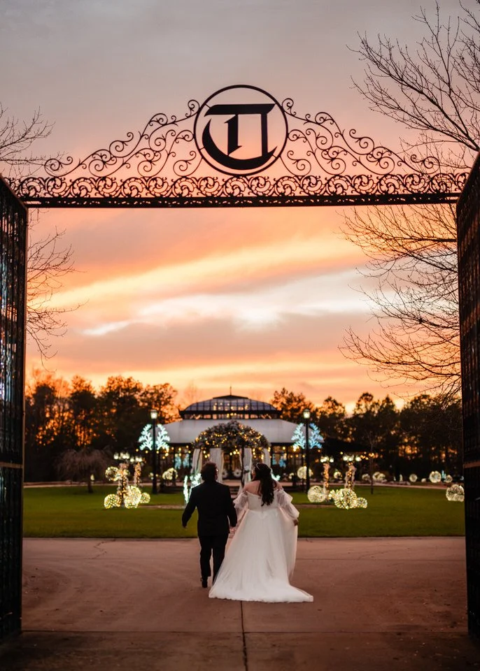 Bride and groom walking hand in hand through Edinburgh West’s ornate iron gates toward the illuminated venue beneath an orange and pink sunset sky.