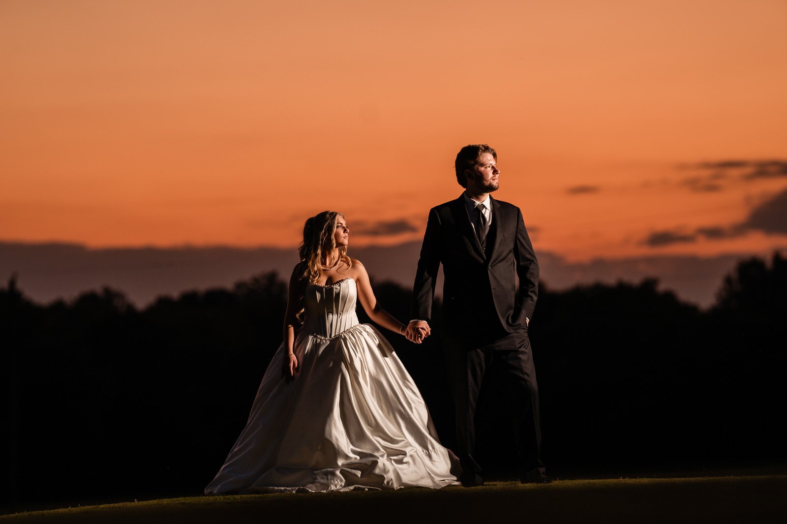 Bride and groom holding hands on a grassy rise at The Barn at Sitton Hill, dramatically lit against a vivid orange sunset and dark tree line.