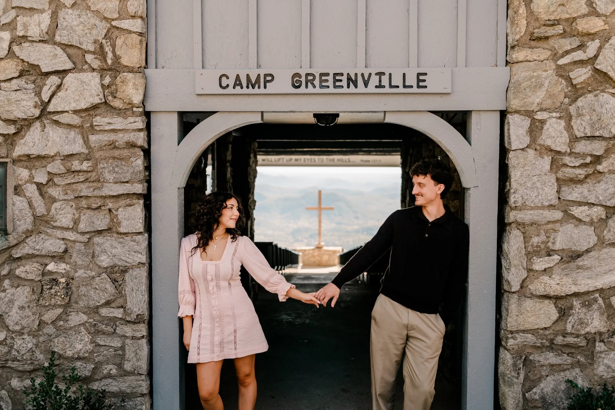 Engaged couple holding hands beneath the Camp Greenville sign at Pretty Place Chapel’s stone entrance, with the cross and mountains visible behind them.