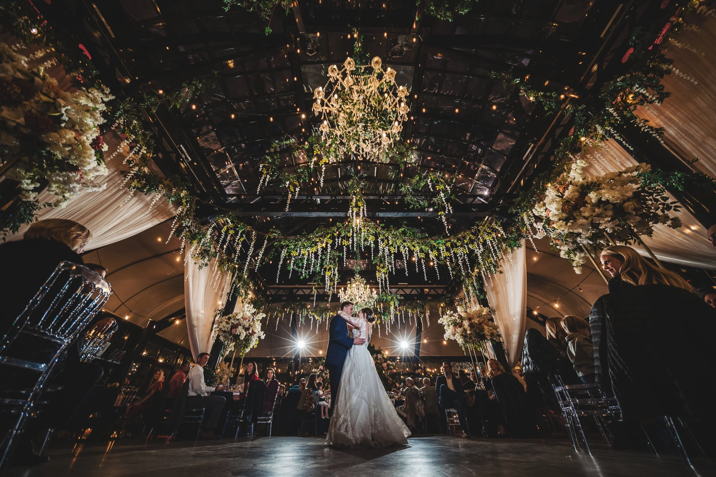 Bride and groom holding hands beneath an ornate iron gazebo at Edinburgh West at sunset, with her long lace train spread across the foreground.