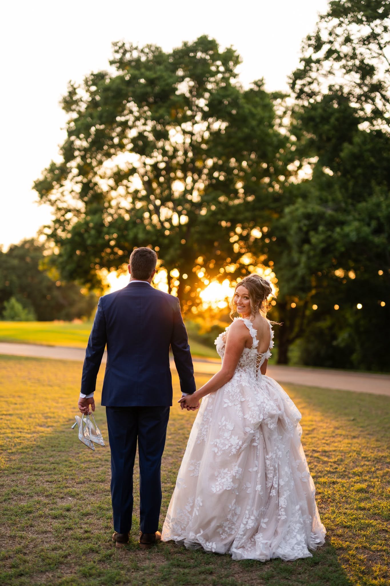 Bride and groom holding hands while walking across the lawn at Edinburgh West at golden hour, with the bride smiling back and the groom carrying her shoes.