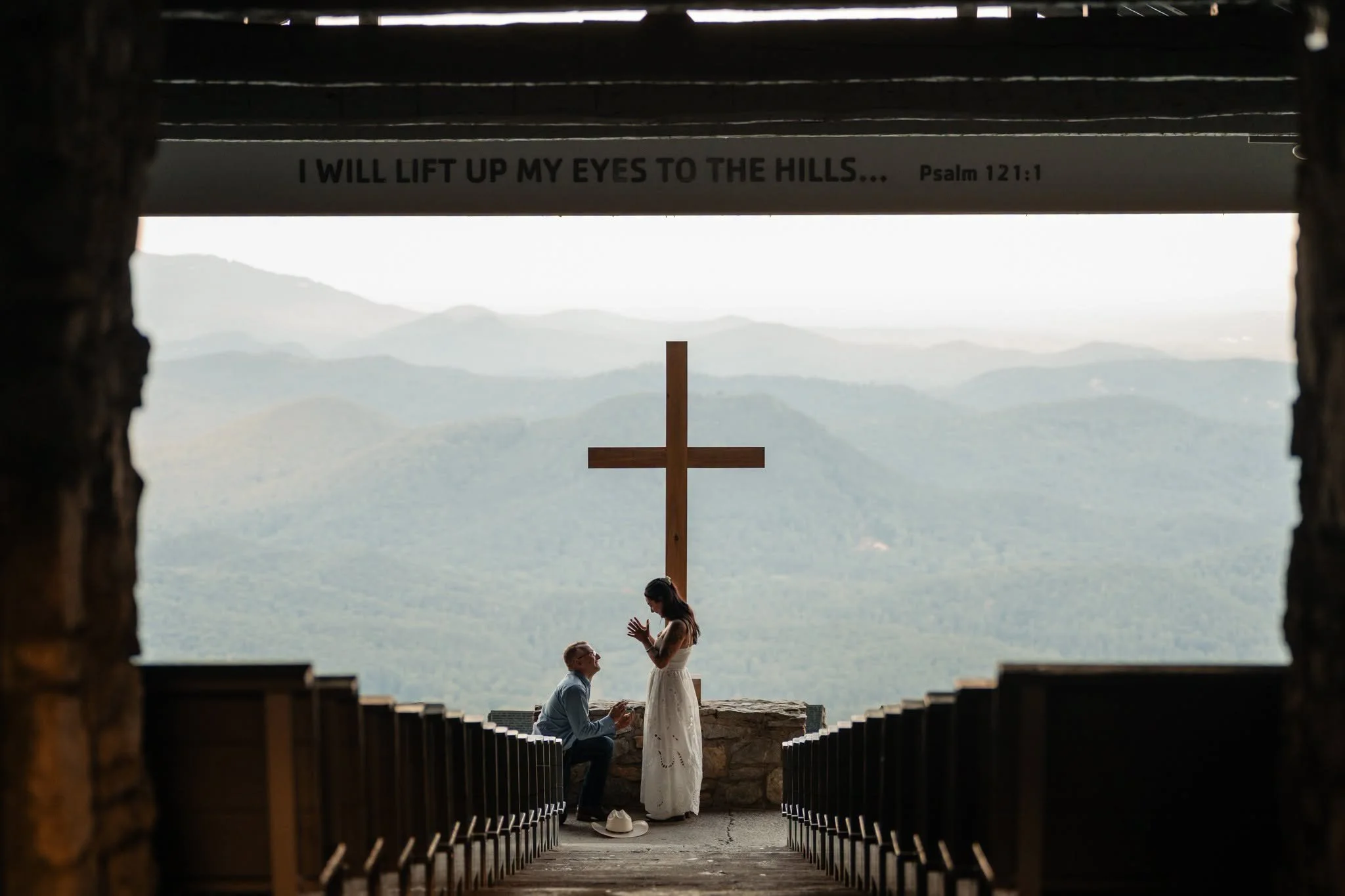 Man kneeling to propose to a woman beneath the cross at Pretty Place Chapel, framed by rows of wooden pews and mountain views.