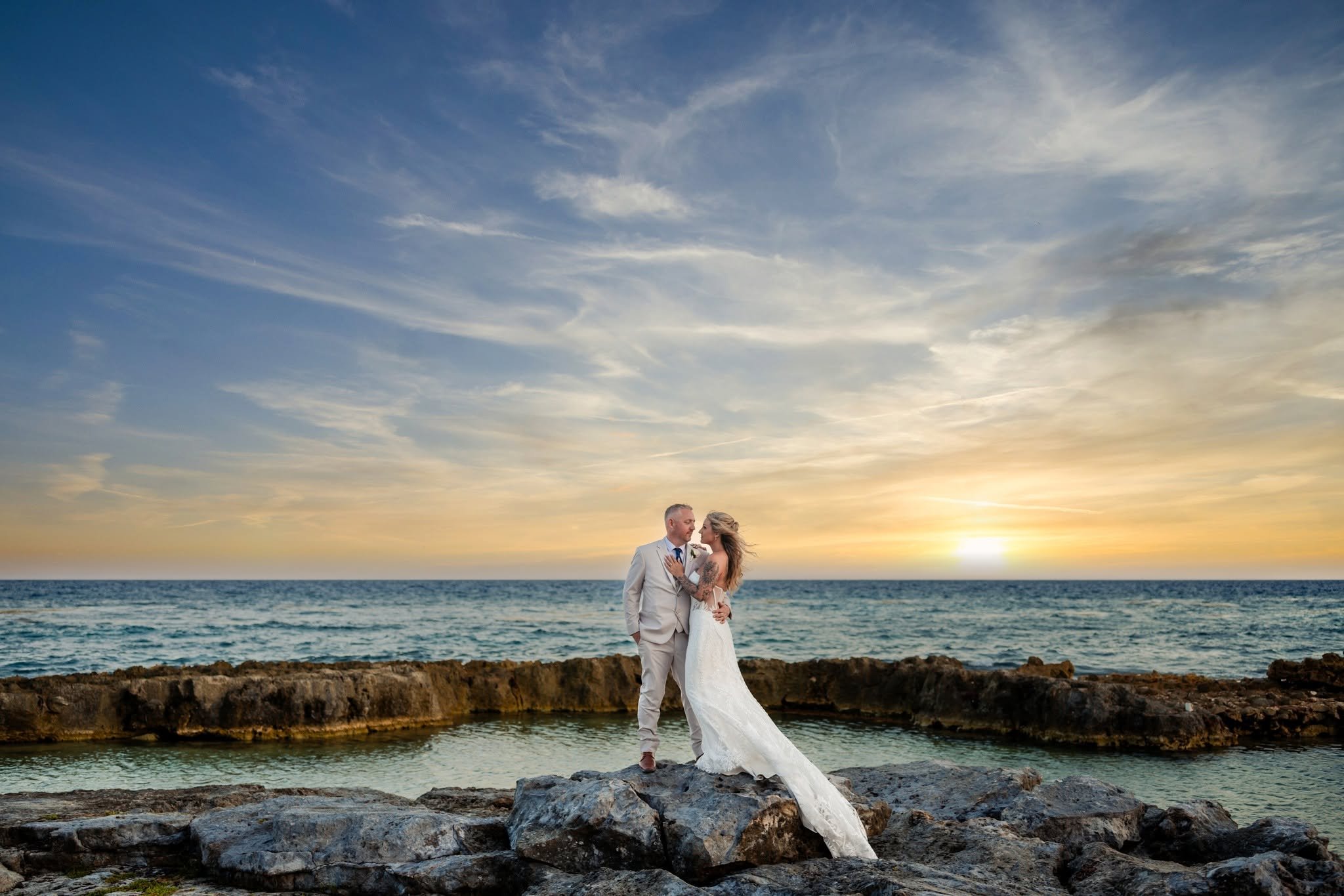 Bride and groom embracing on a rocky shoreline in Jamaica, with ocean waves and a golden sunset behind them.
