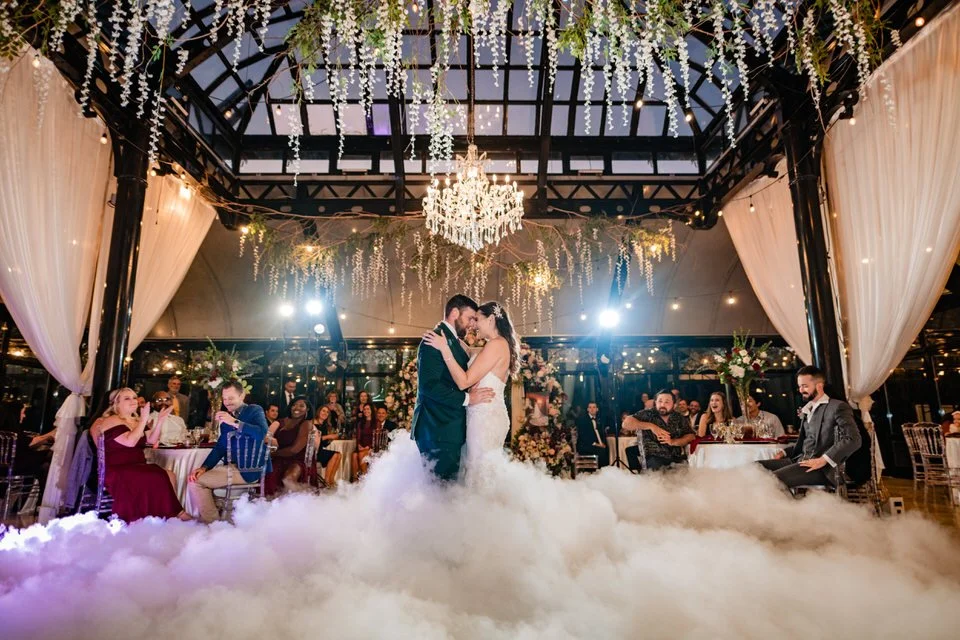 Bride and groom sharing their first dance amid low-lying fog beneath a crystal chandelier and hanging flowers in Edinburgh West’s glass conservatory, surrounded by wedding guests.