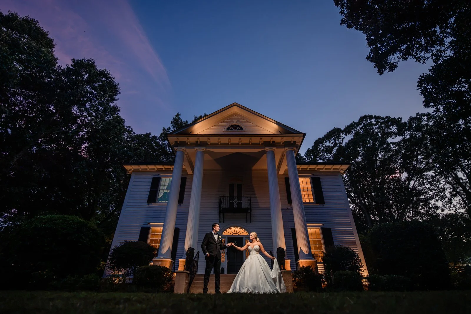 Bride and groom holding hands in front of Duncan Estate’s illuminated historic house at twilight beneath a blue and pink sky in Spartanburg, SC.