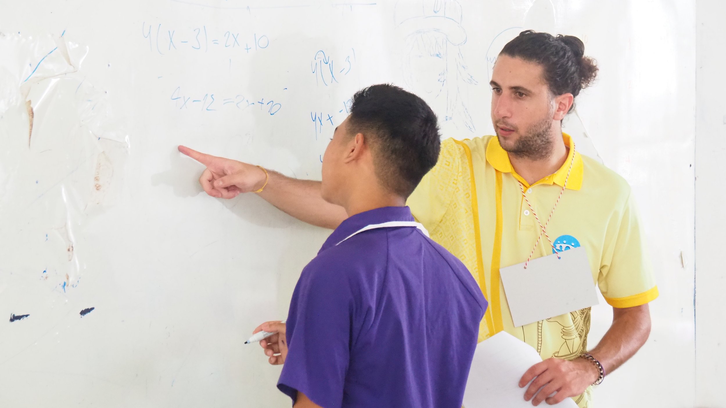 One student, one in a purple shirt and the other a Kids English Thailand teacher in a yellow shirt, standing in front of a whiteboard with mathematical equations and drawings, engaging in a conversation.