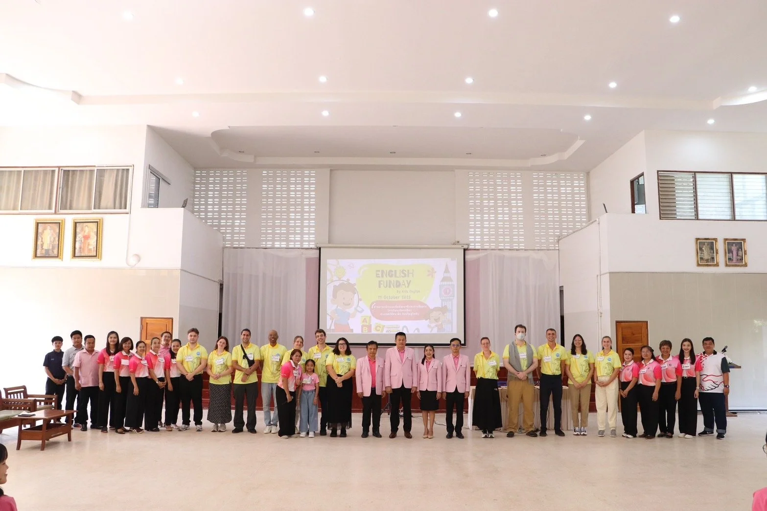 Group photo of about 25  Kids English Thailand teachers standing in a row inside a spacious hall with a large screen behind them displaying 'English Funday' and cartoon characters.