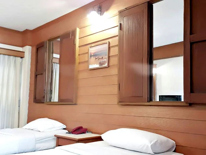 A hotel room with two single beds, wooden window shutters, and a small nightstand. A picture hangs on the wall, and curtains are drawn to the side of the window.