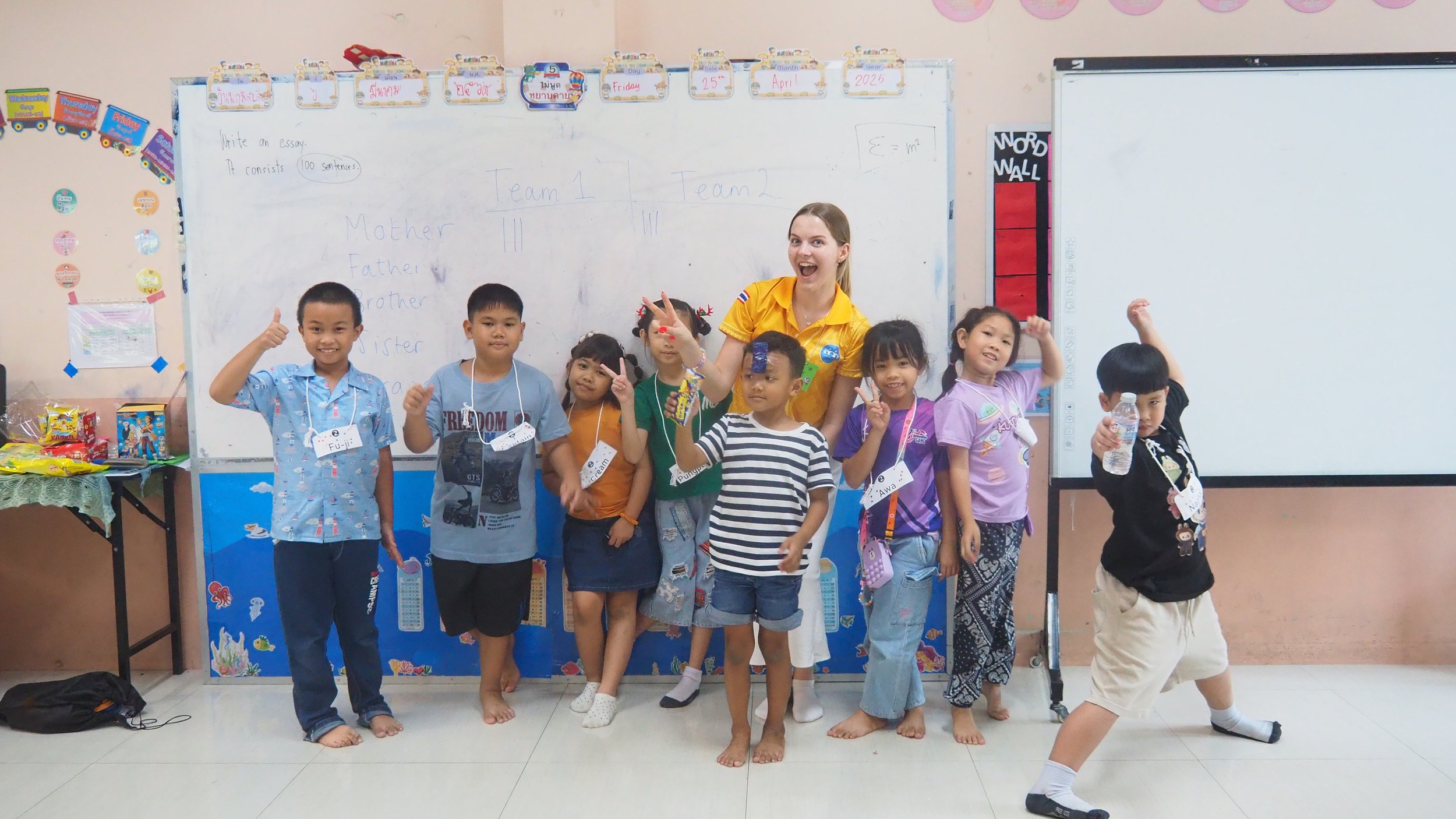 Group of Kids English Thailand children and a Kids English Thailand teacher posing in a classroom, some children giving thumbs up, others making peace signs, with whiteboard and colorful decorations in the background.