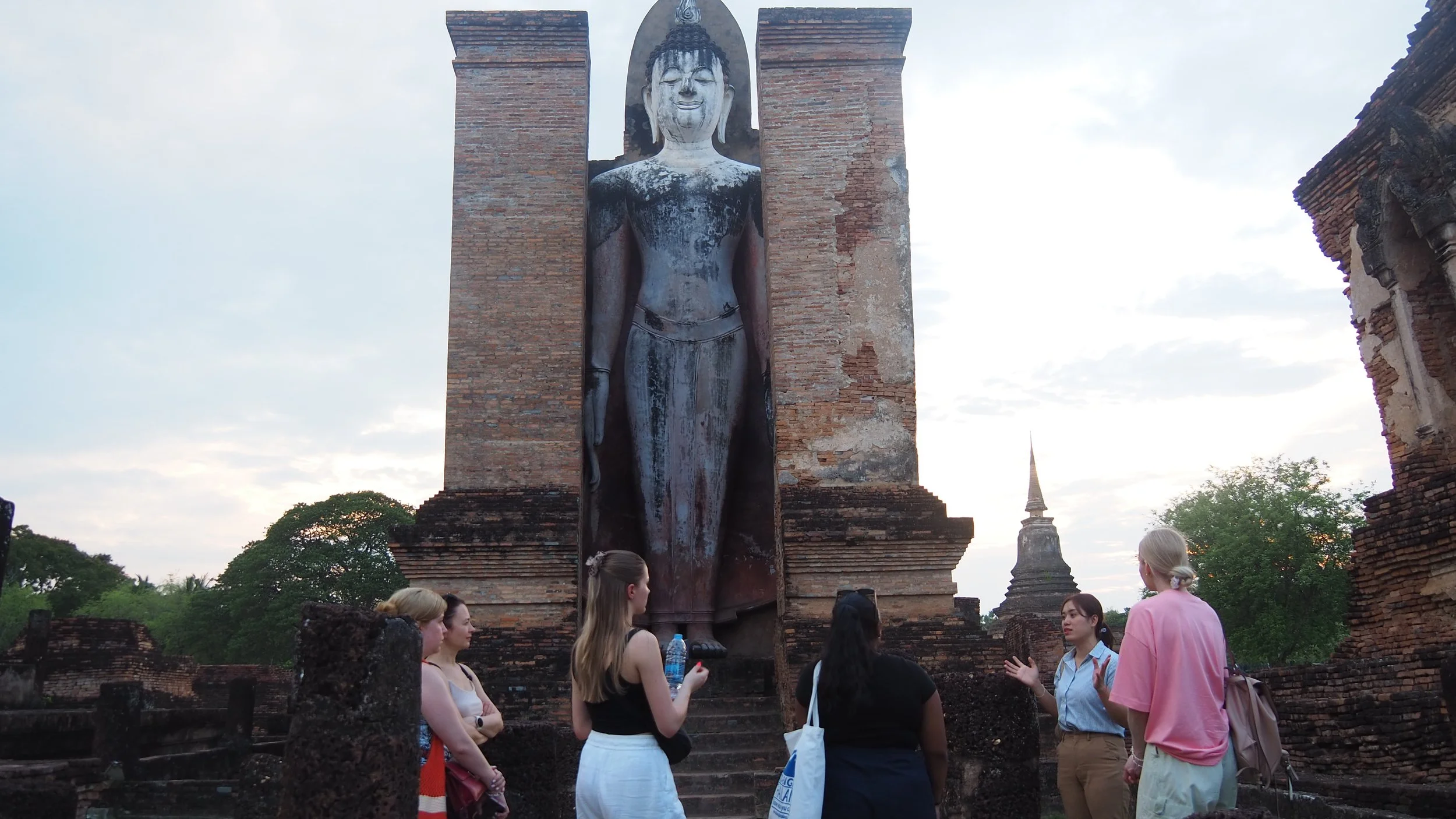 A group of Kids English Thailand teachers stands at ancient brick ruins with a large, weathered Buddha image carved into a wall behind them, during daytime with a cloudy sky.