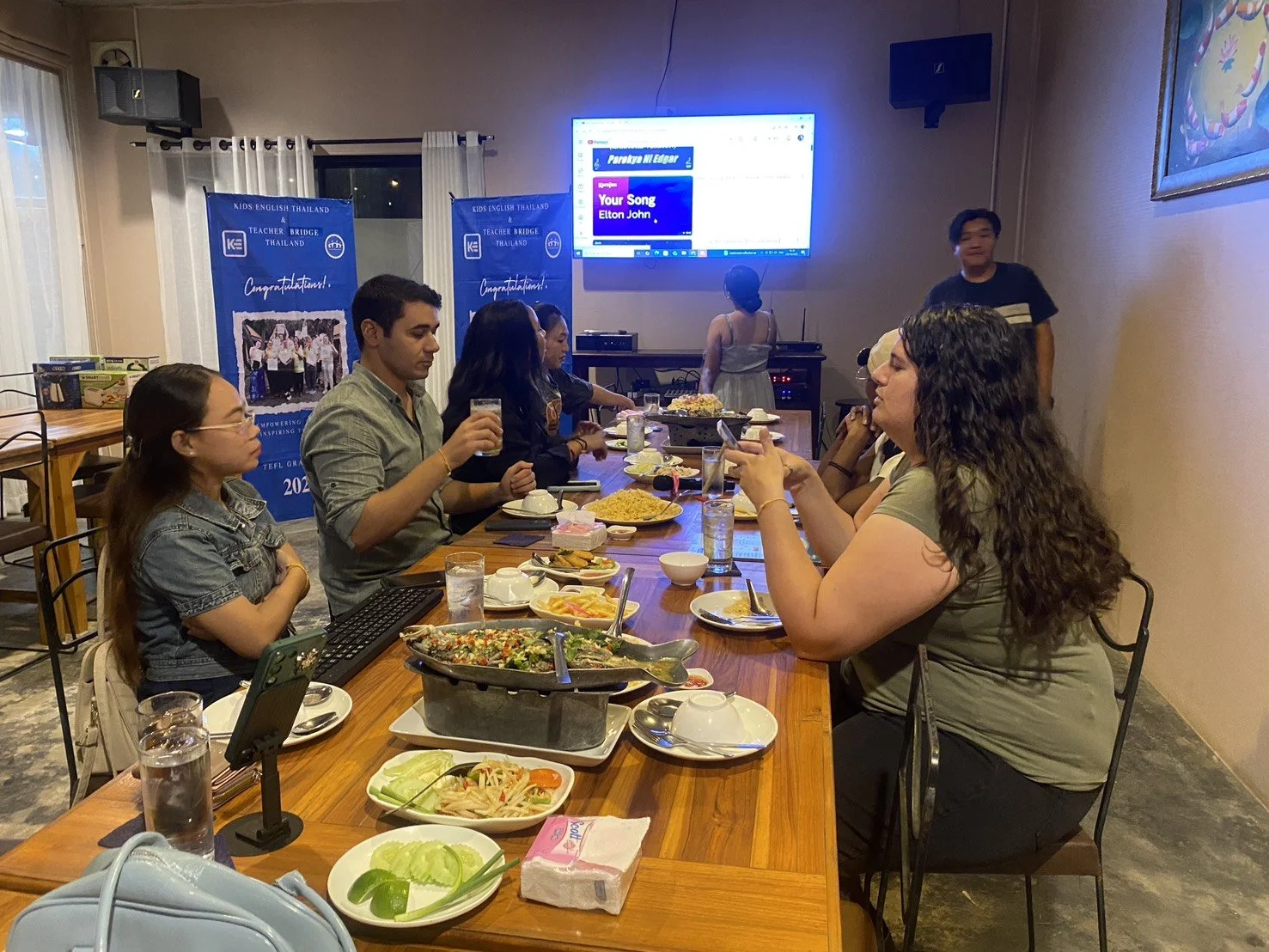 People sitting at a dinner table with food, drinks, and plates, in a room with banners and a large screen displaying text. Kids English Thailand experience!