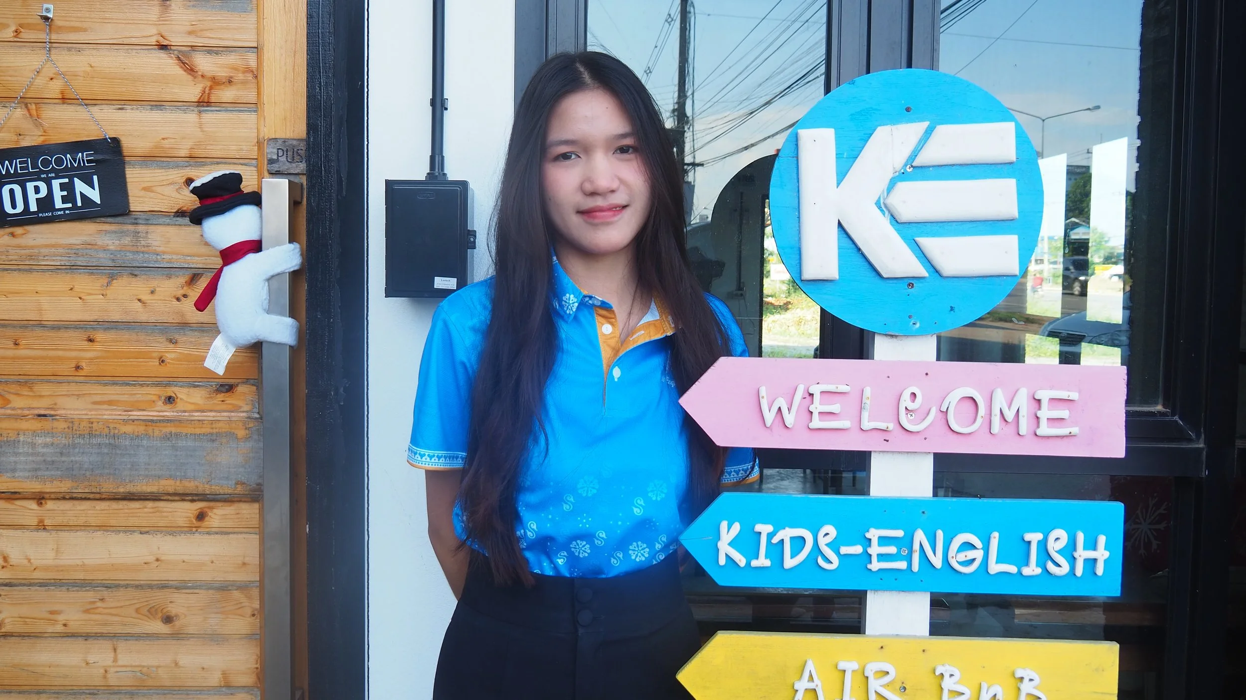 A woman in a blue shirt holding a pink 'Welcome' sign, standing next to a colorful sign with directions for kids' English classes outside a building with wooden siding and glass windows.