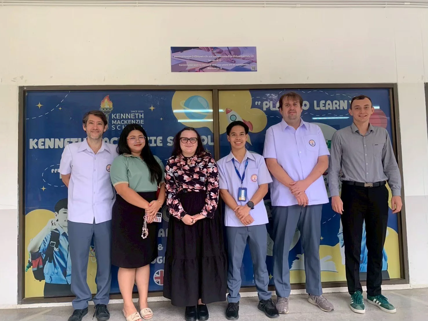 Group of six people from Kids English Thailand standing in front of a school banner, smiling for the photo.