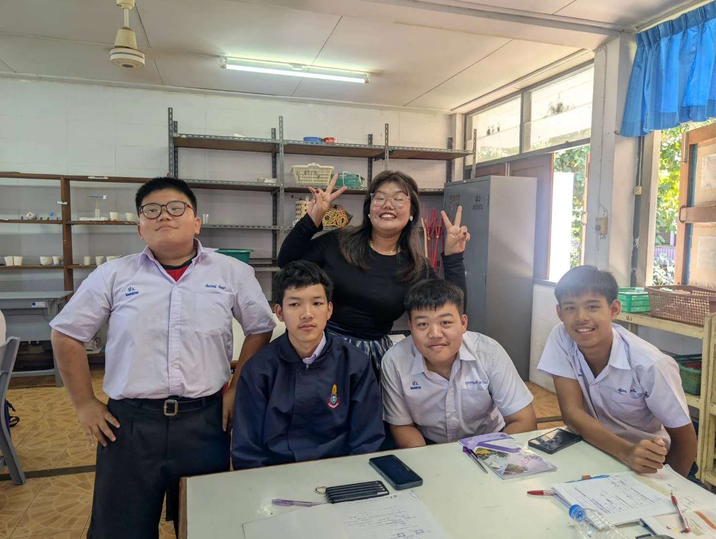 A group of four boys and one Kids English Thailand teacher , posing inside a classroom. The KET teacher is standing behind the boys, making a peace sign with both hands, while the boys are sitting or standing at a table with notebooks and phones. 