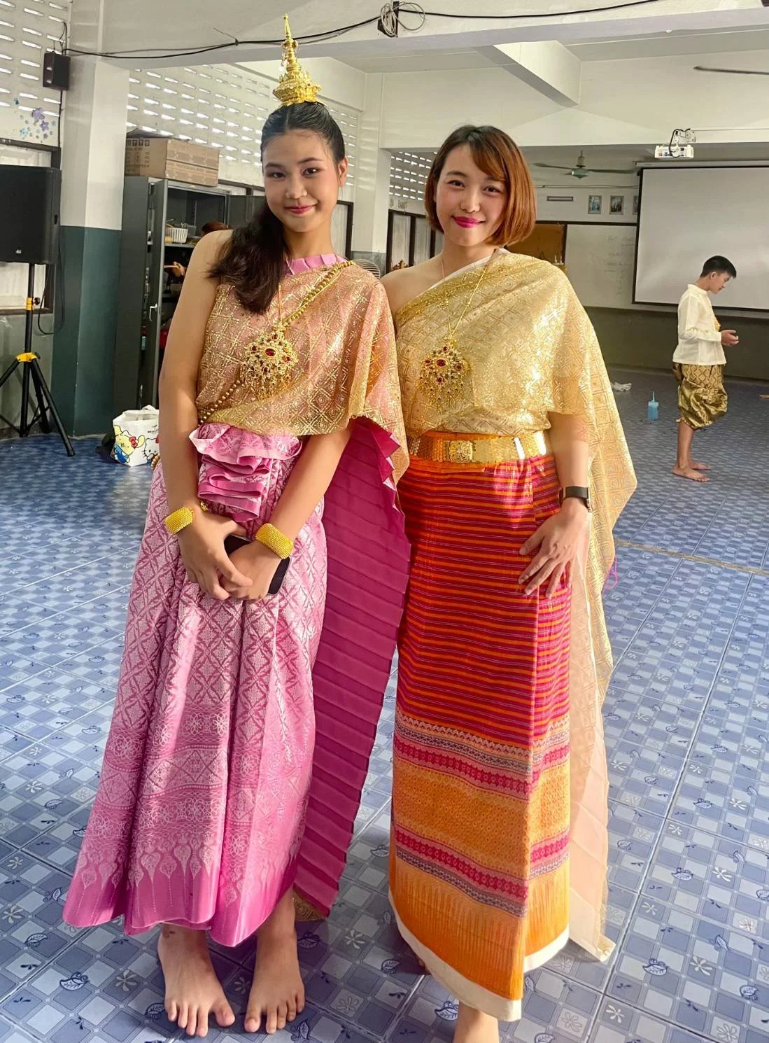 Two young Kids English Thailand teachers wearing traditional Thai clothing and gold jewelry, standing indoors on a tiled floor. They are smiling and posing for the photo, with a boy in traditional Thai attire in the background.
