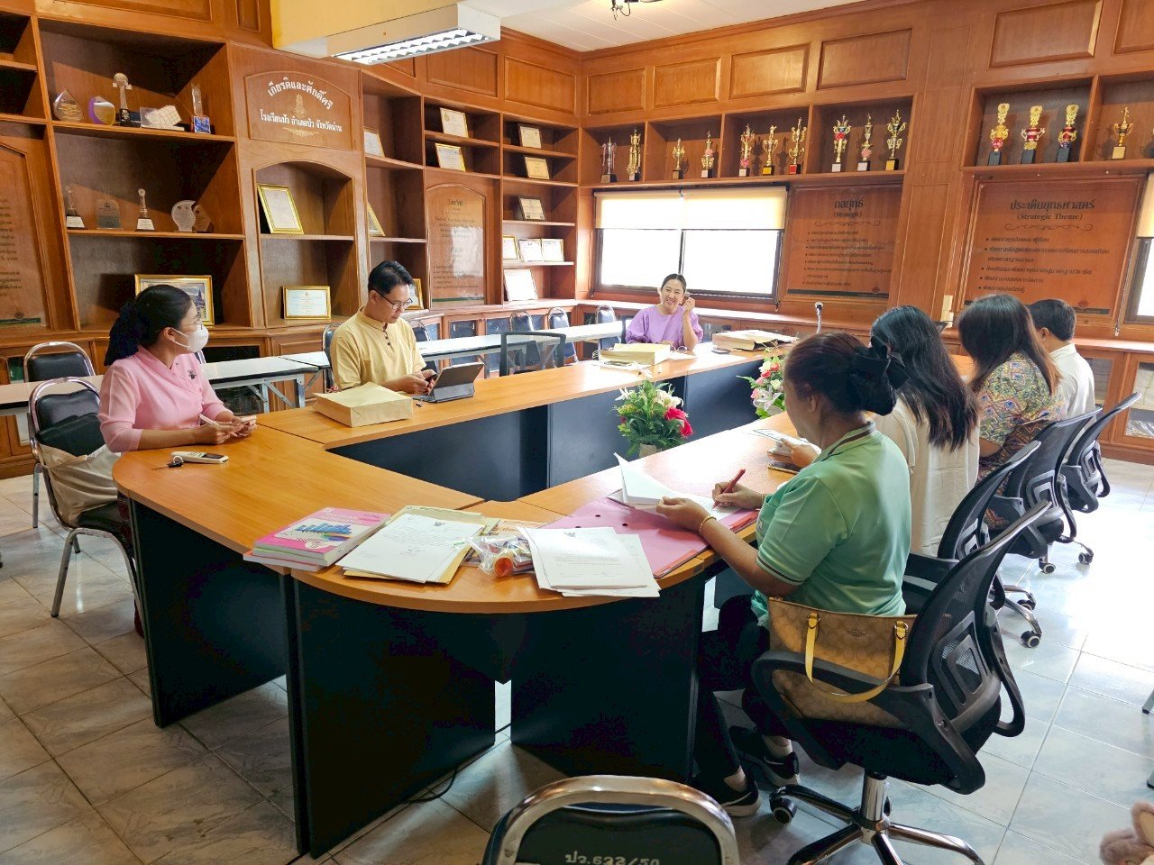 A group of people from Kids English Thailand in a meeting room with wooden walls, sitting around a U-shaped table, some taking notes and others listening to a woman speaking at the end of the table. 