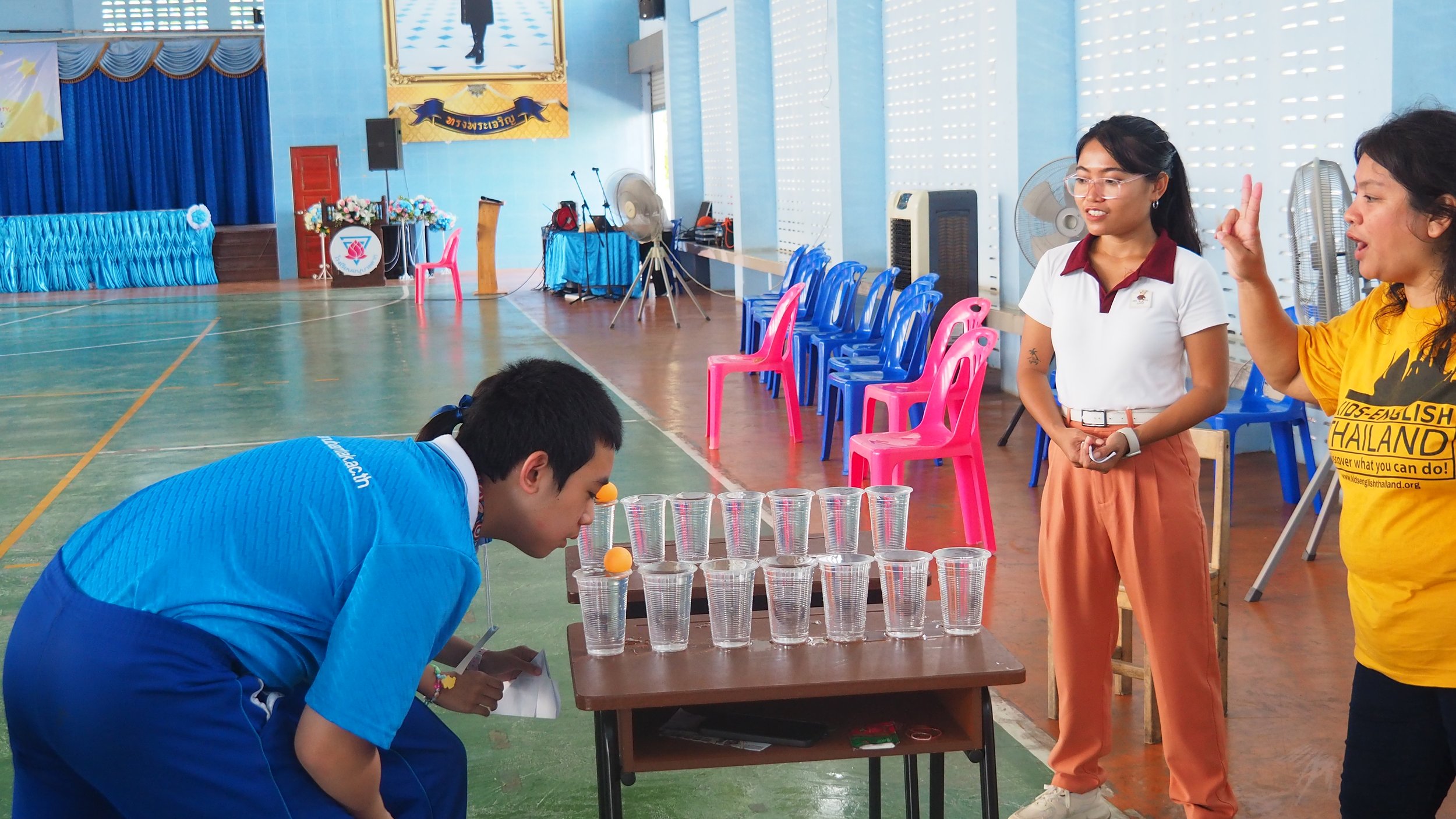 A boy in blue uniform with a clown nose is leaning over a table with cups and ping pong balls, participating in a game. Two Kids English Thailand teachers are standing nearby, one with glasses and another wearing a yellow KET shirt.