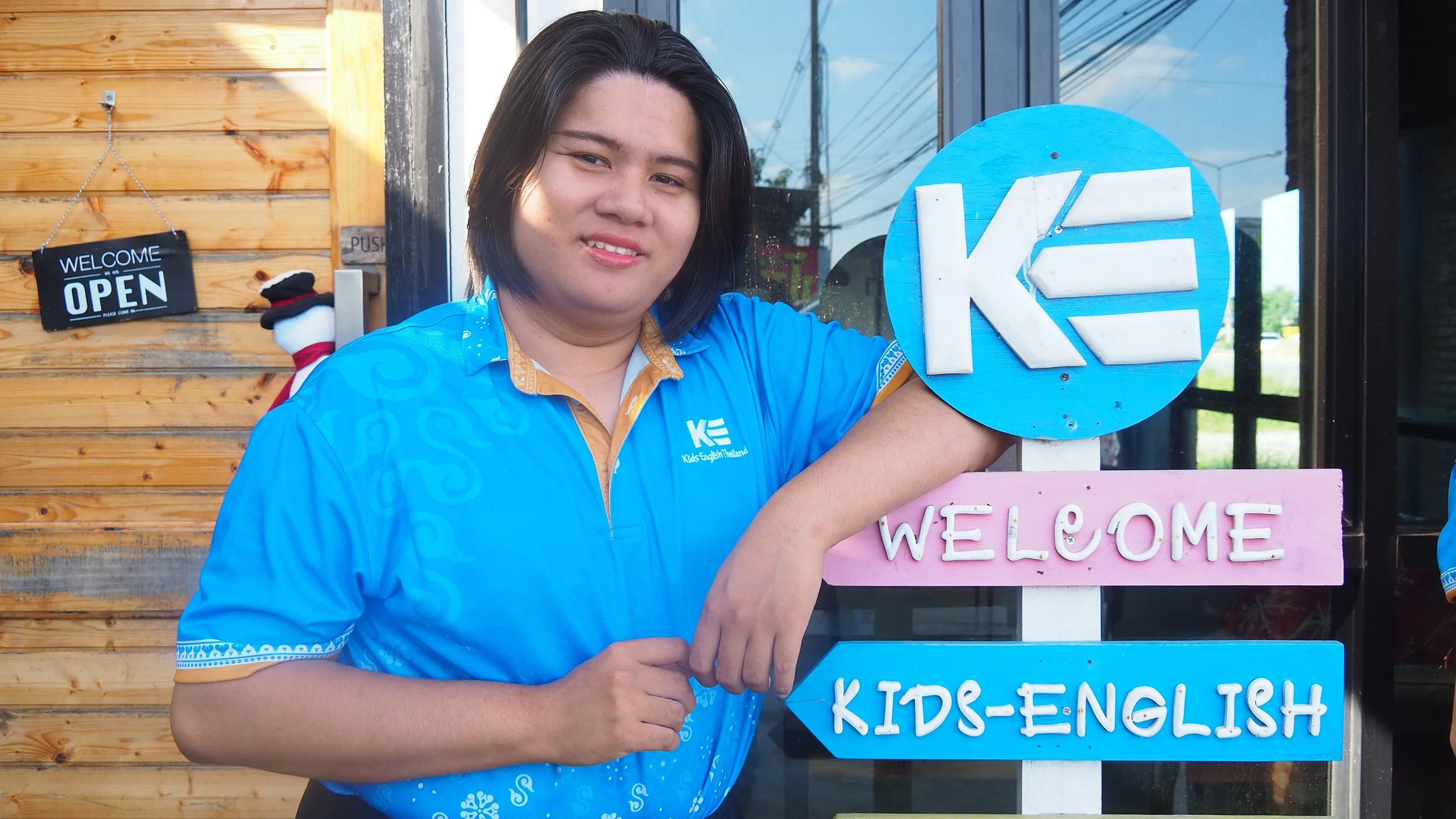 A woman standing next to colorful signs that say "WELCOME" and "KIDS-ENGLISH" outside a building with a wooden wall and glass windows.