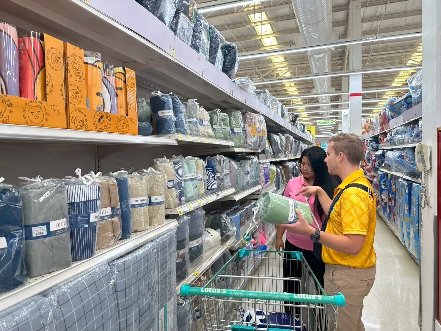 Two people from Kids English Thailand shopping in a home goods aisle, looking at bedding and comforters, with aisle shelves filled with various packaged linens and textiles.