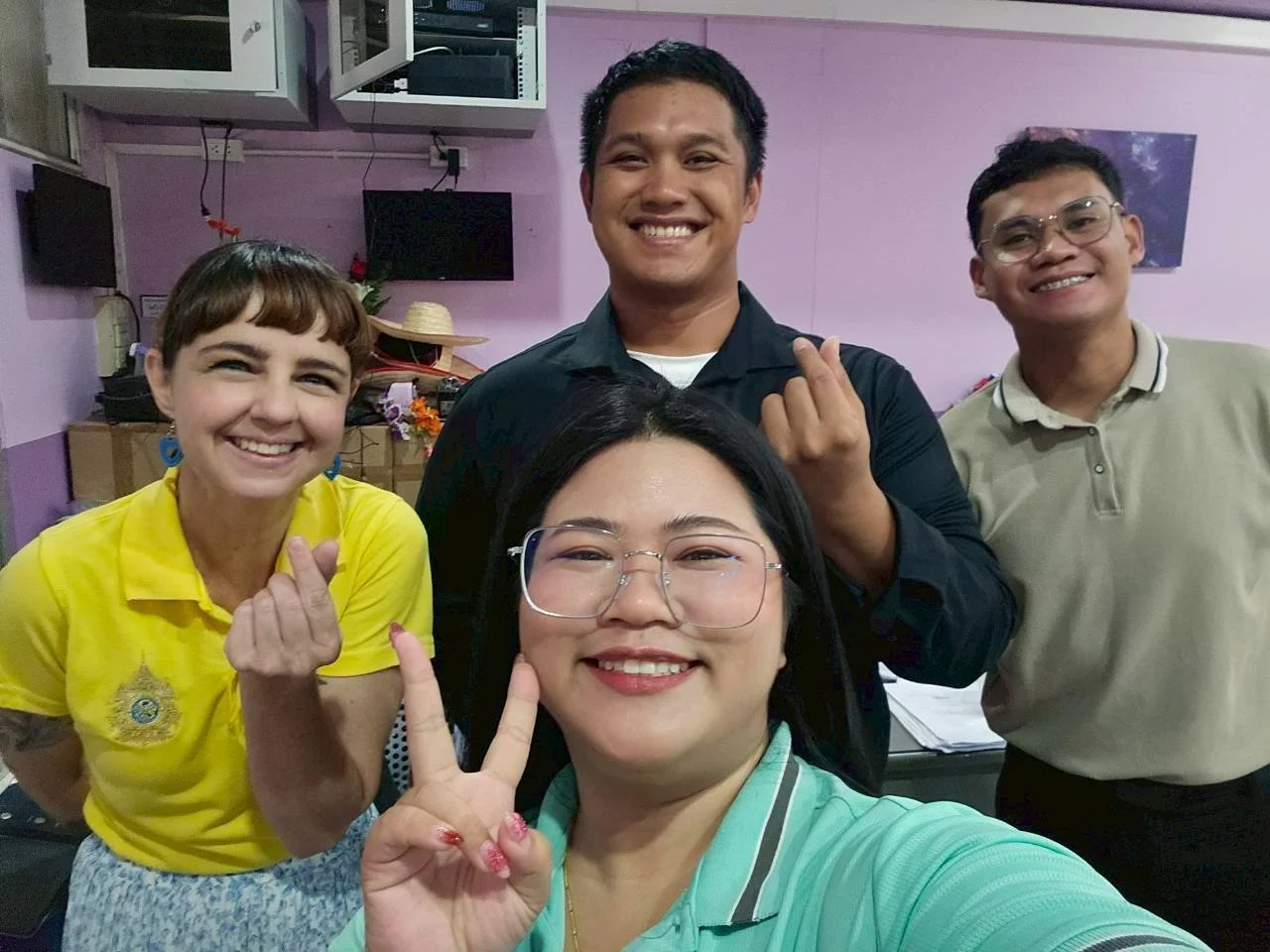 Group of five smiling people from Kids English Thailand taking a selfie indoors, some making finger heart gestures. Four are standing in the background, and one woman is in the foreground wearing glasses and giving a peace sign.
