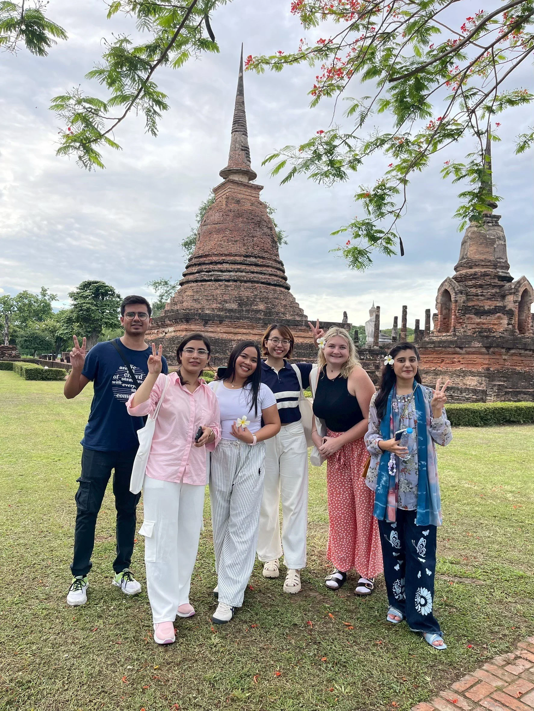 Group of six Kids English Thailand teachers and volunteers and interns posing outdoors in front of ancient brick spires, with green grass and trees, under a cloudy sky, some making peace signs.