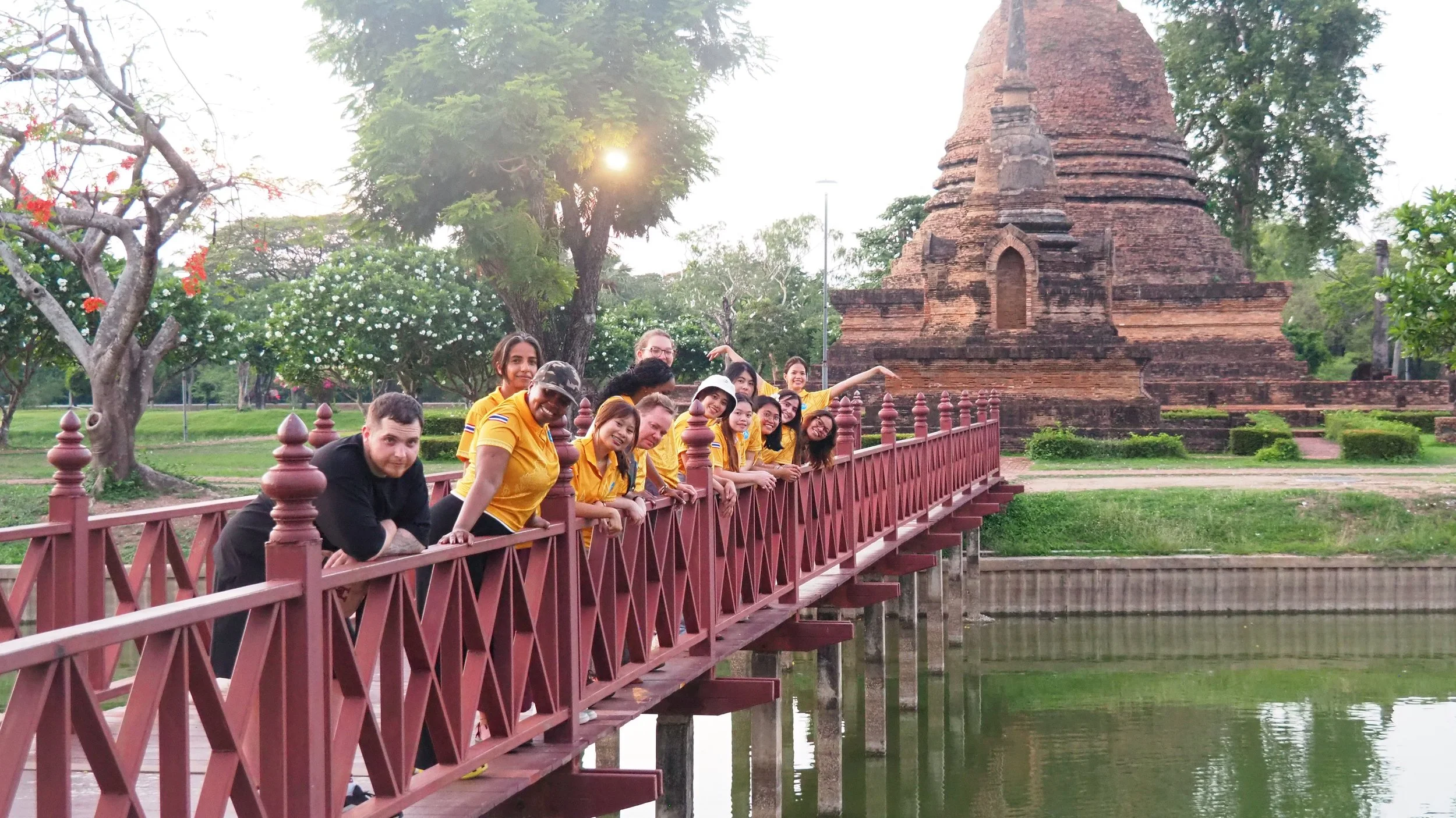 A group of Kids English Thailand teachers standing on a pinkish-red bridge over a pond in front of ancient brick pagoda, with trees and greenery in the background.