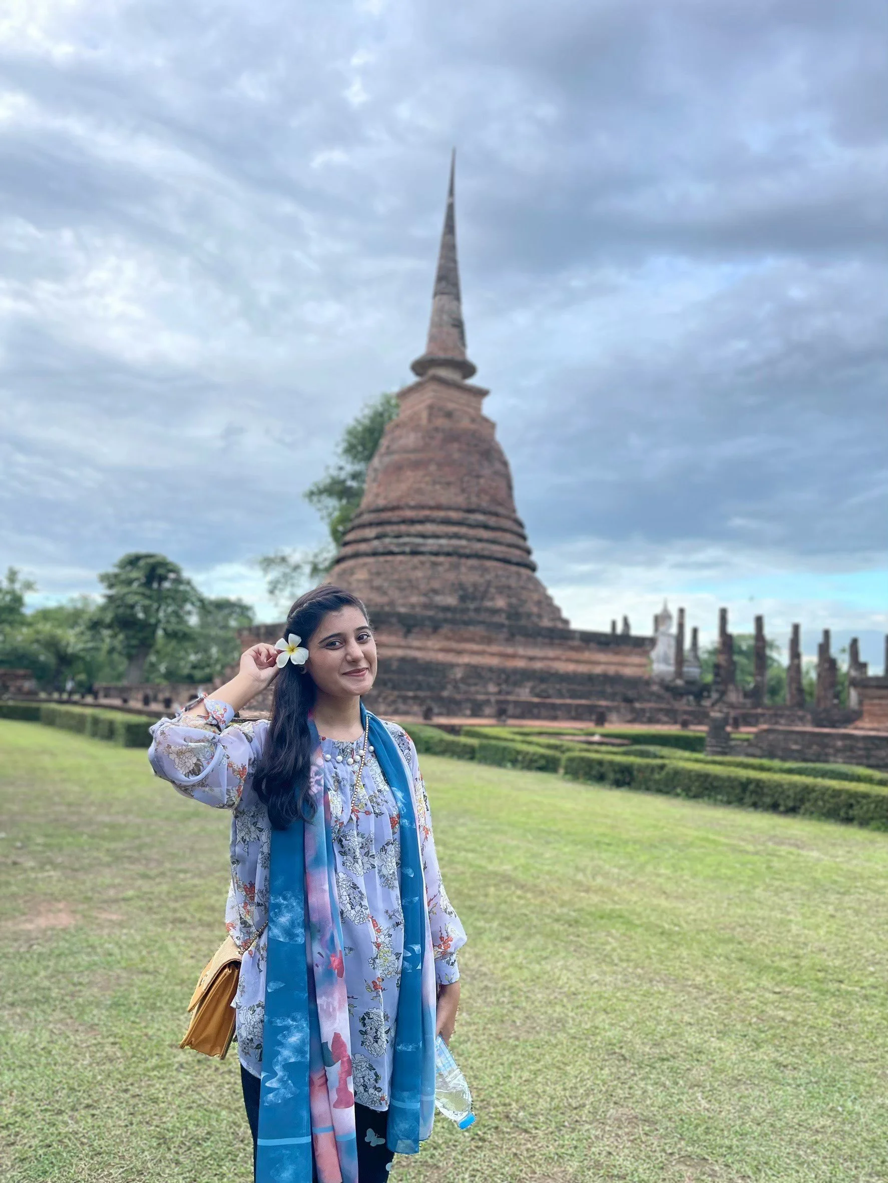 A Kids English Thailand teacher  with a white flower in her hair, wearing a floral blouse and a colorful scarf, smiling at the camera while holding a face mask in her right hand.