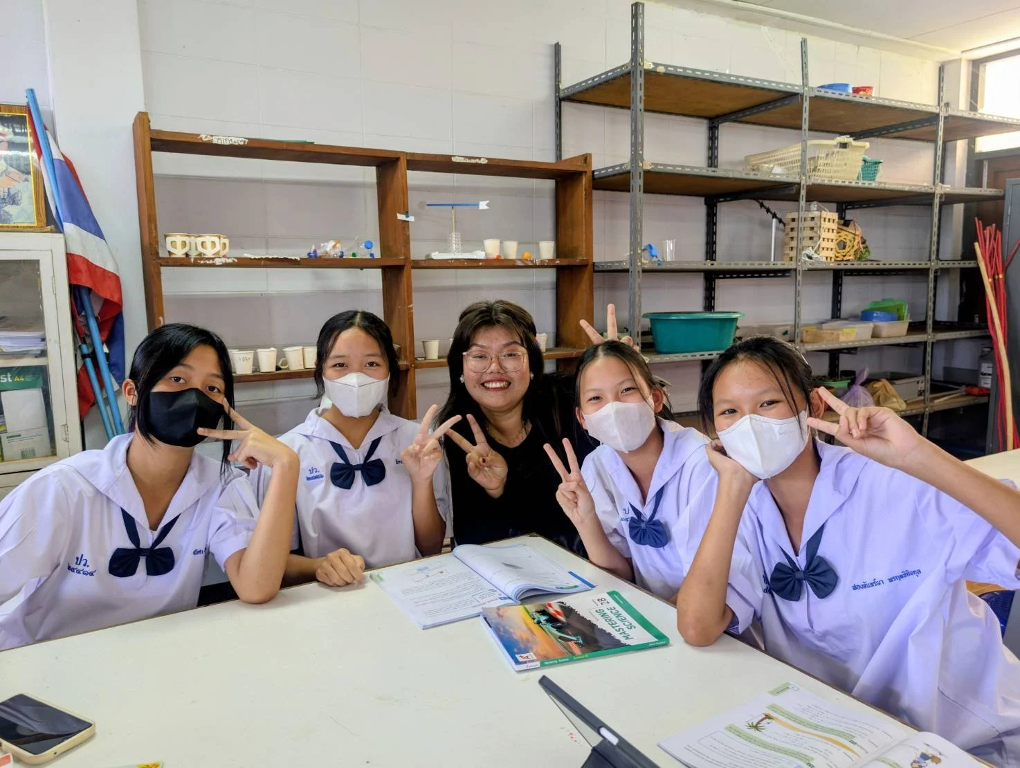 Group of five students and a Kids English Thailand teacher sitting at a table in a classroom, all smiling and making peace signs, wearing white school uniforms and face masks.