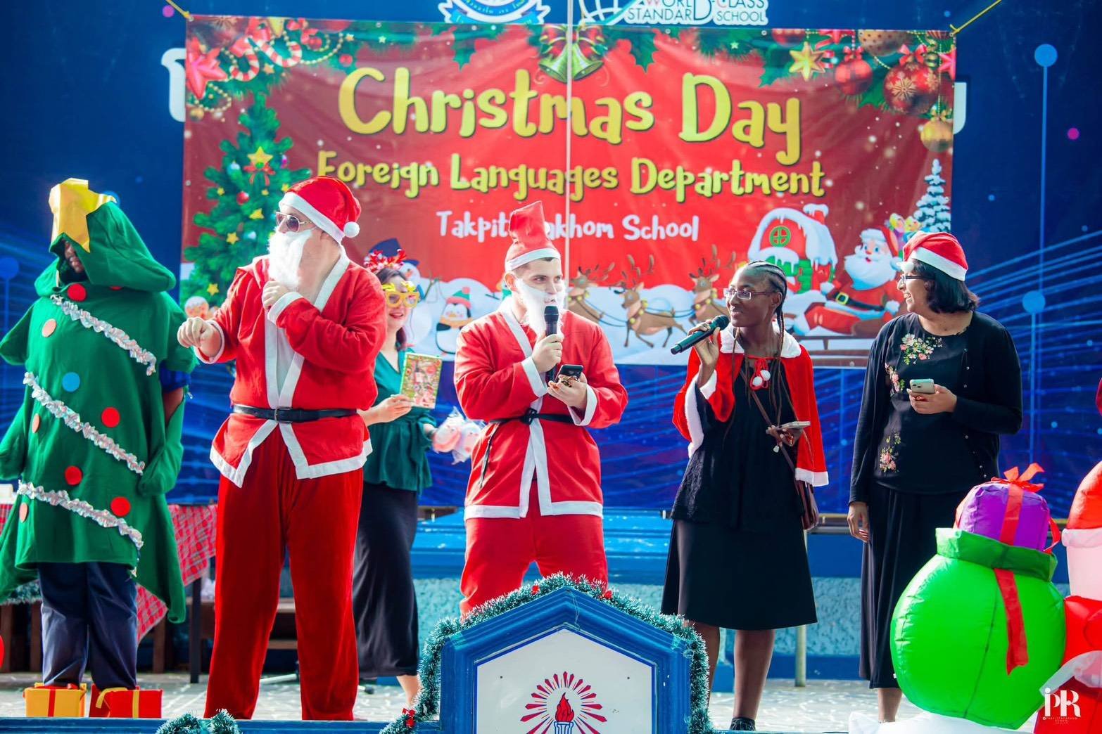 A group of Kids English Thailand teachers dressed in Christmas costumes, including a person dressed as Santa Claus, a Christmas tree costume, and other festive attire, are on stage during a Christmas celebration.
