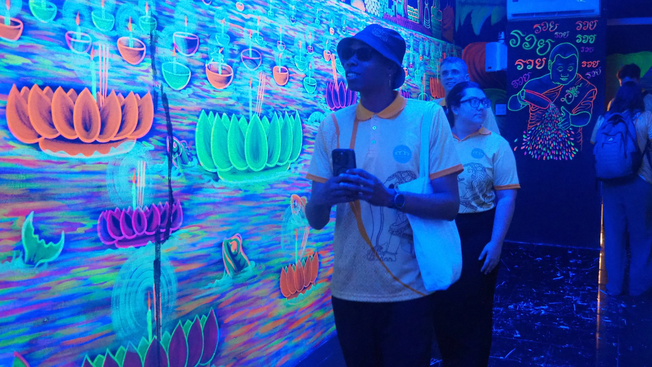Kids English Thailand teachers viewing neon-lit artwork in a dark room, with vibrant lotus flowers and water motifs on the wall.