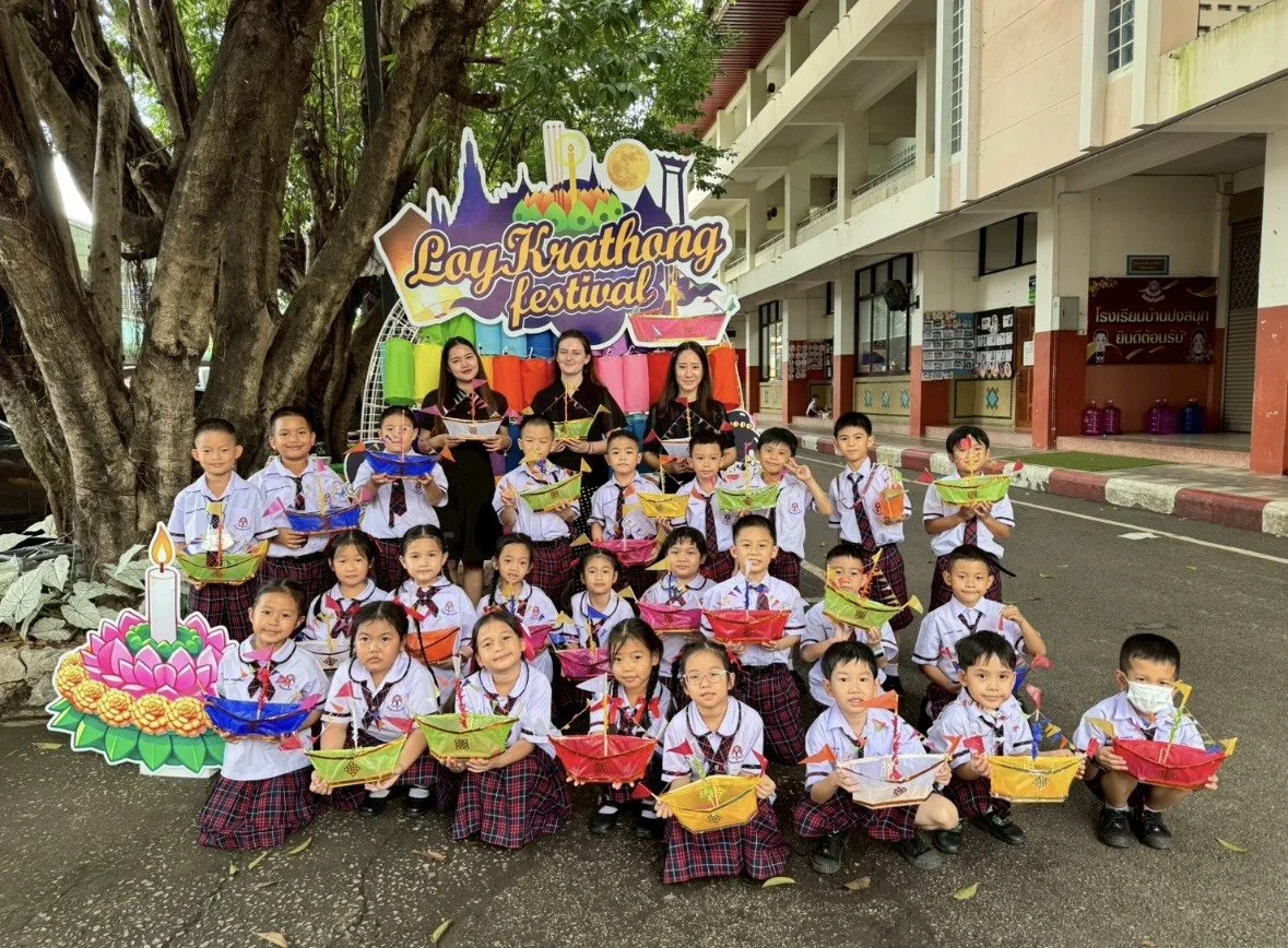 Group of children and Kids English Thailand teachers celebrating Loy Krathong festival outdoors, with a large colorful sign in the background, some children holding decorated krathongs, and a cartoon illustration of a lit candle.