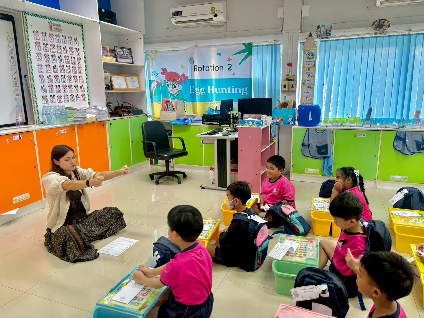 A teacher from Kids English Thailand in a classroom with young students sitting on the floor, engaging in a teaching activity. The classroom has colorful cabinets, a large chart on the wall, and a banner that reads 'Rotation 2 Egg Hunting.' 