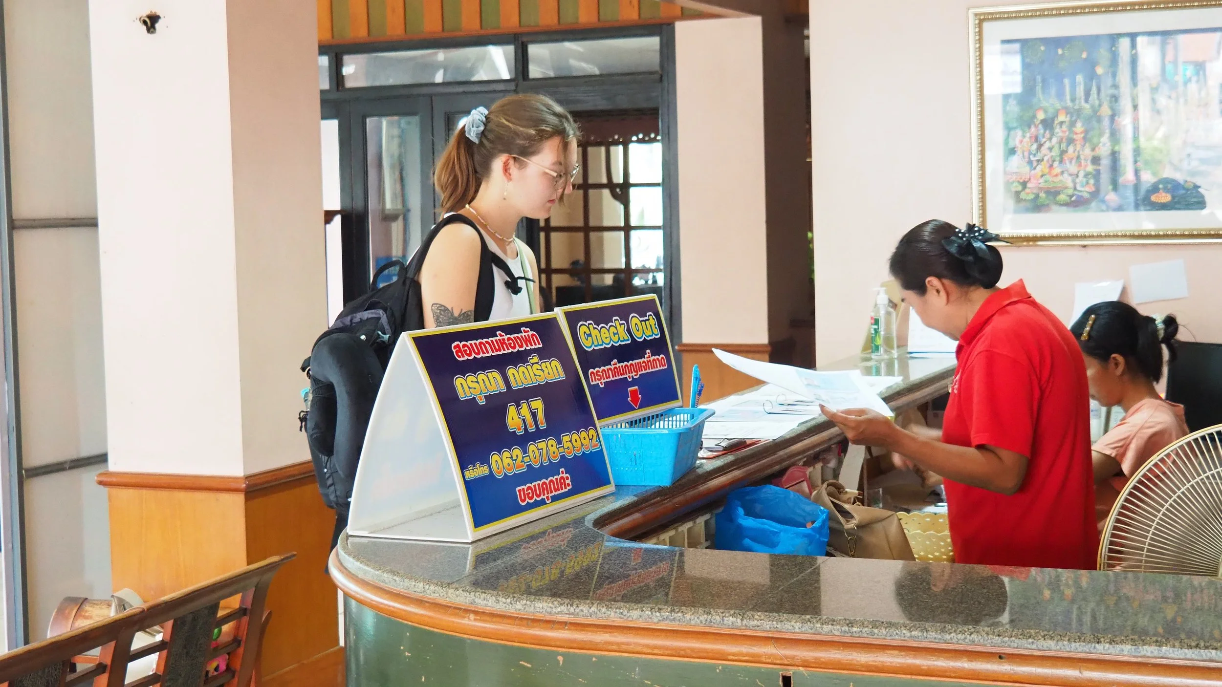 A Kids English Thailand teacher with glasses and a backpack checking out at a hotel reception desk, where a staff member in a red uniform is reading a document. Two guests are seated nearby.