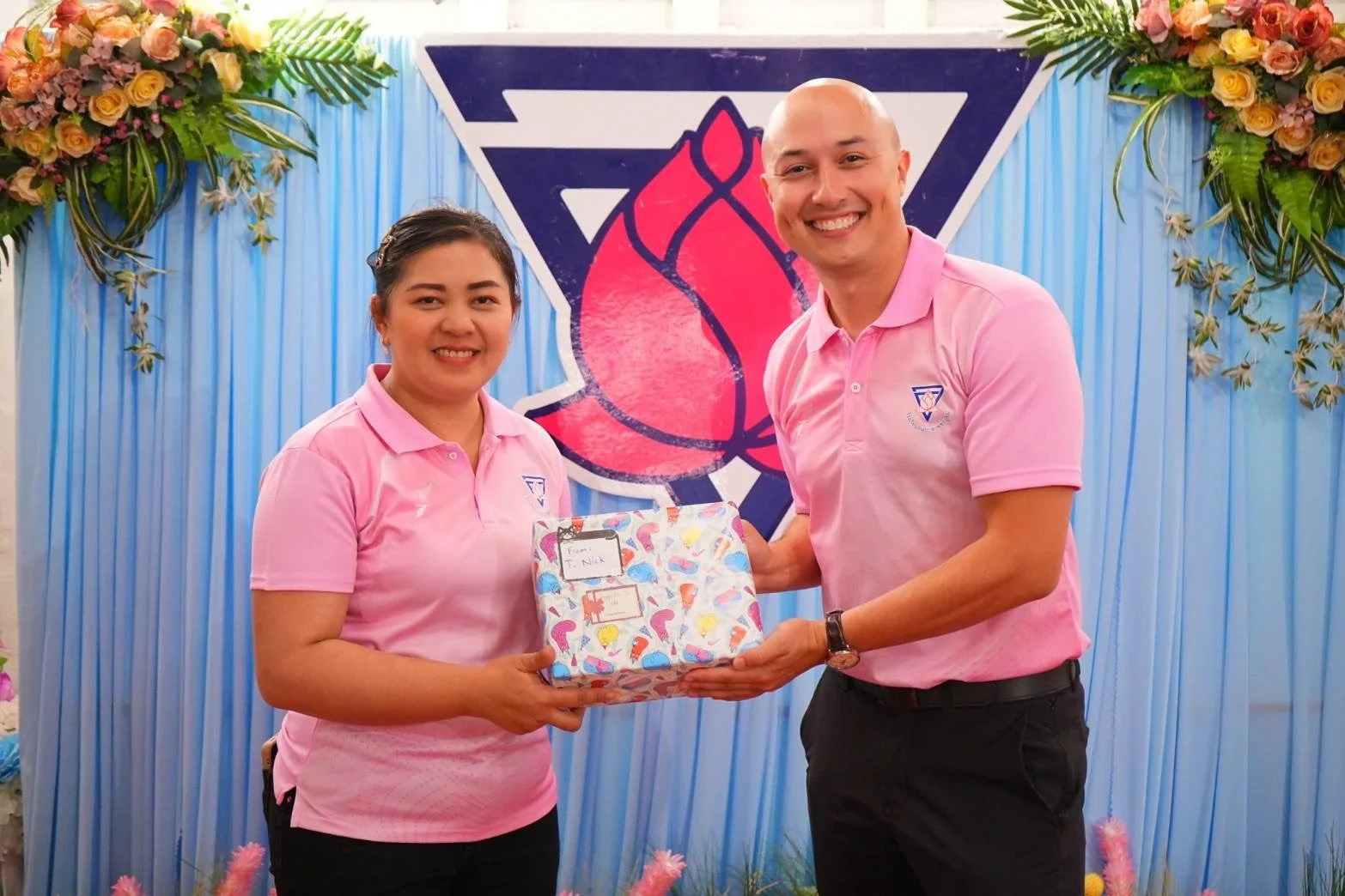 A man and woman from Kids English Thailand in pink shirts exchanging a gift at a celebration event, with blue and floral decorations and a large logo featuring a heart and a triangle in the background.