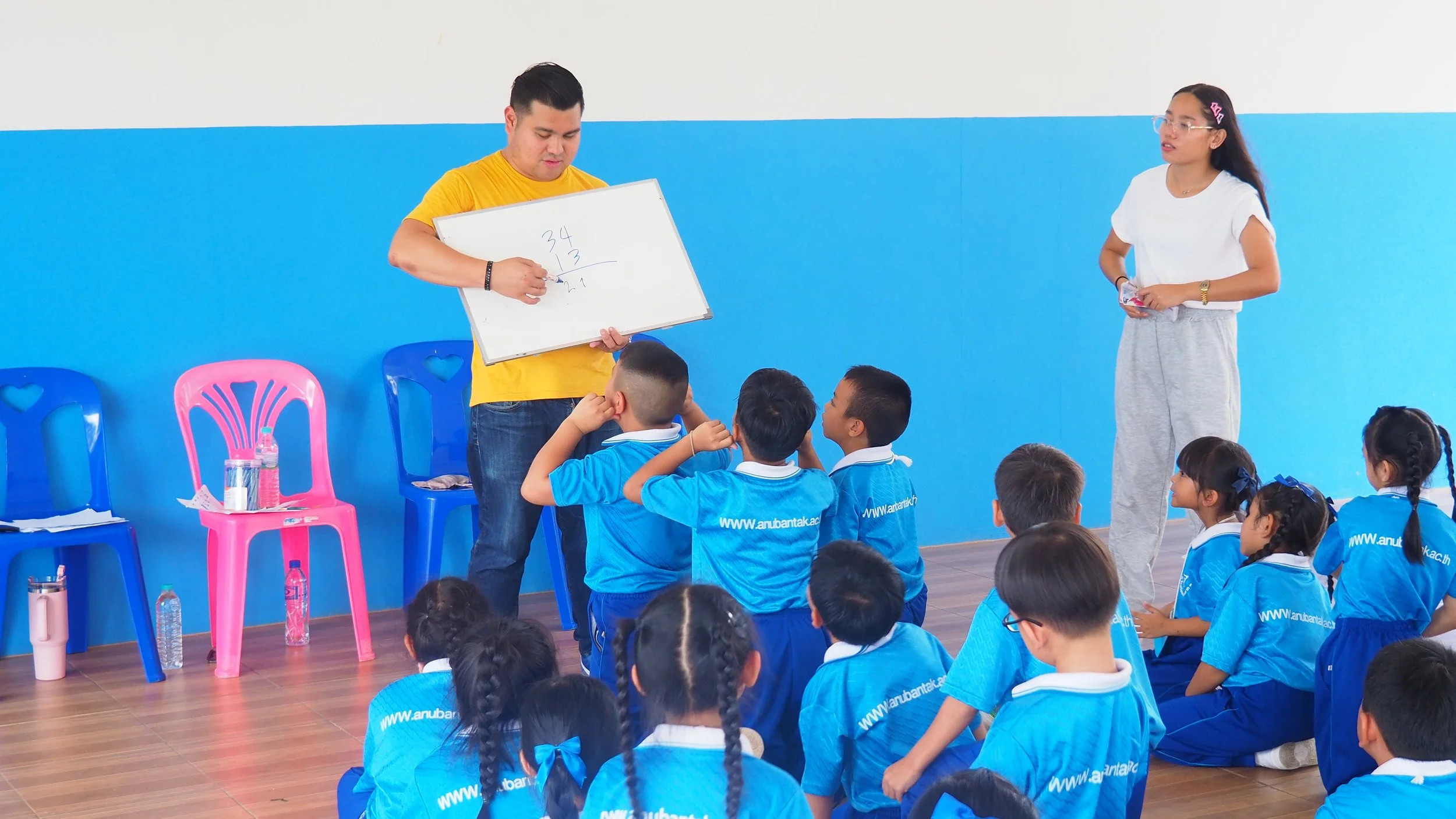 A classroom with young students in matching blue uniforms sitting on the floor and a Kids English Thailand teacher at the front. A male Kids English Thailand teacher in a yellow KET shirt is writing math problems on a whiteboard.