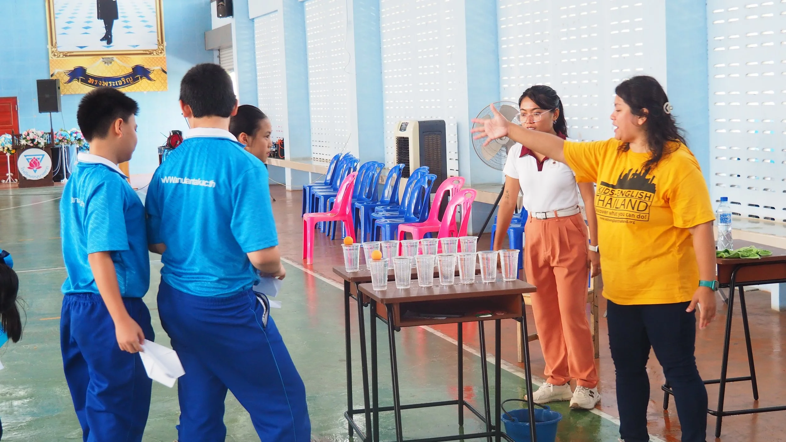 Two children in blue school uniforms receiving instructions from two Kids English Thailand teachers in a gymnasium, with a table of clear cups and small orange balls.