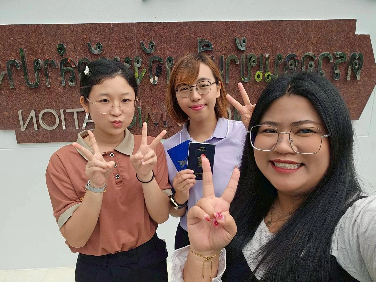 Three women from Kids English Thailand taking a selfie in front of a sign that has text in a non-Latin script. They are smiling and making peace signs.