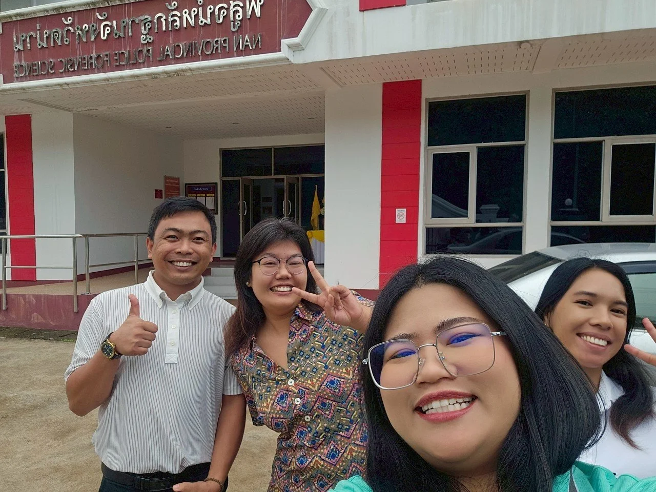 Four people from Kids English Thailand smiling and taking a selfie in front of a building with a sign in a language with characters similar to Thai, featuring white walls with red accents, and a parked white car.