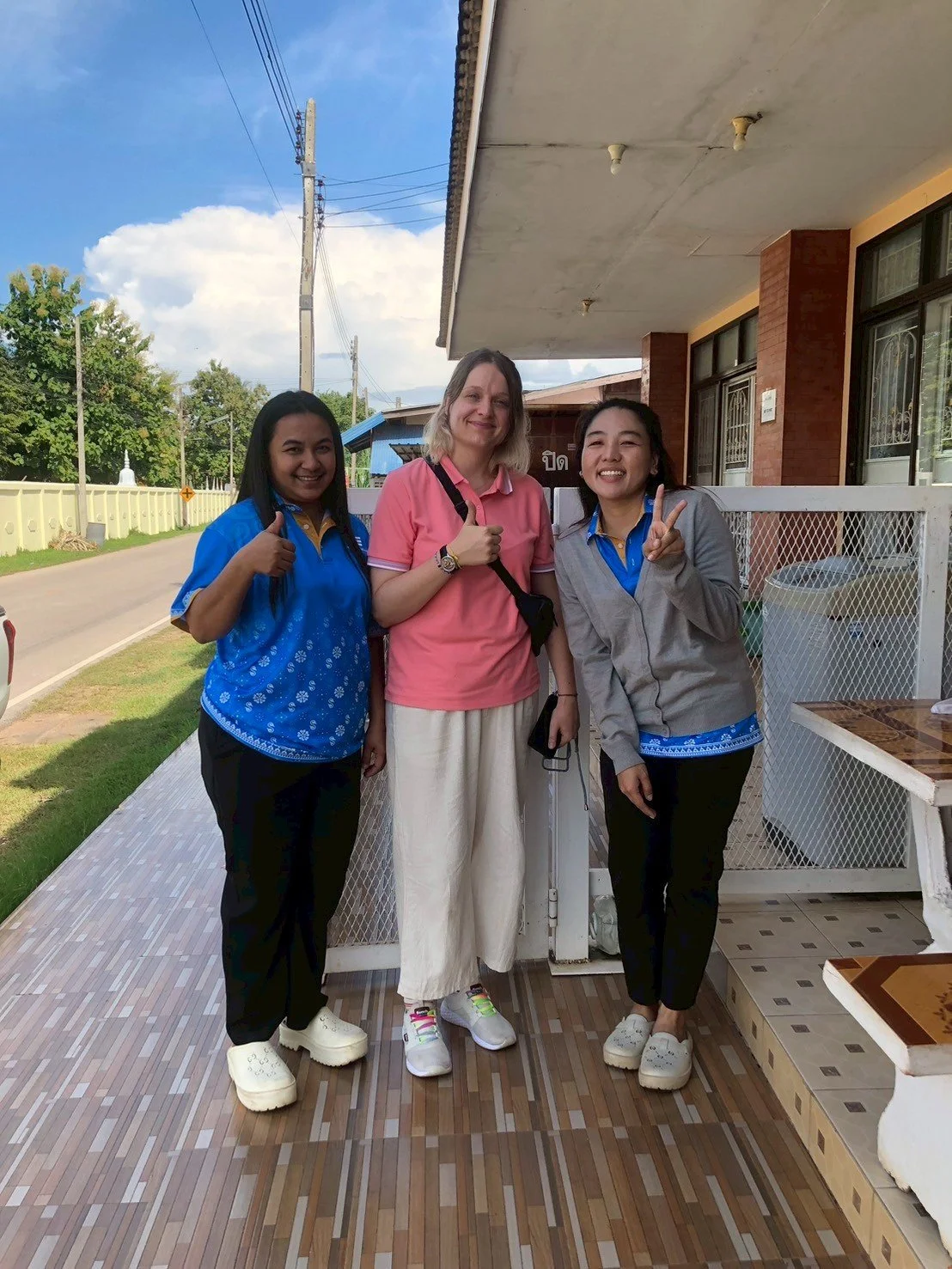 Three women from Kids English Thailand standing together outdoors on a sidewalk, smiling and making peace signs. The woman in the middle is wearing a pink shirt and white pants, while the women on either side are in blue uniforms. 