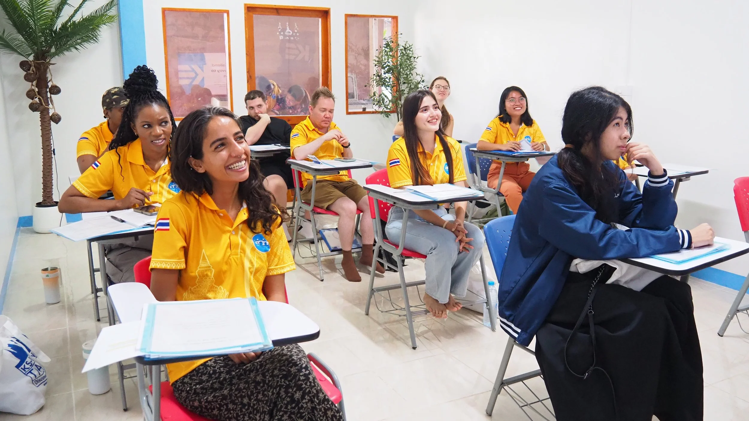 A classroom with diverse Kids English Thailand teachers, some smiling and attentive, some focused, seated at desks with notebooks and papers, with a large plant on the left and conference-style windows at the back.