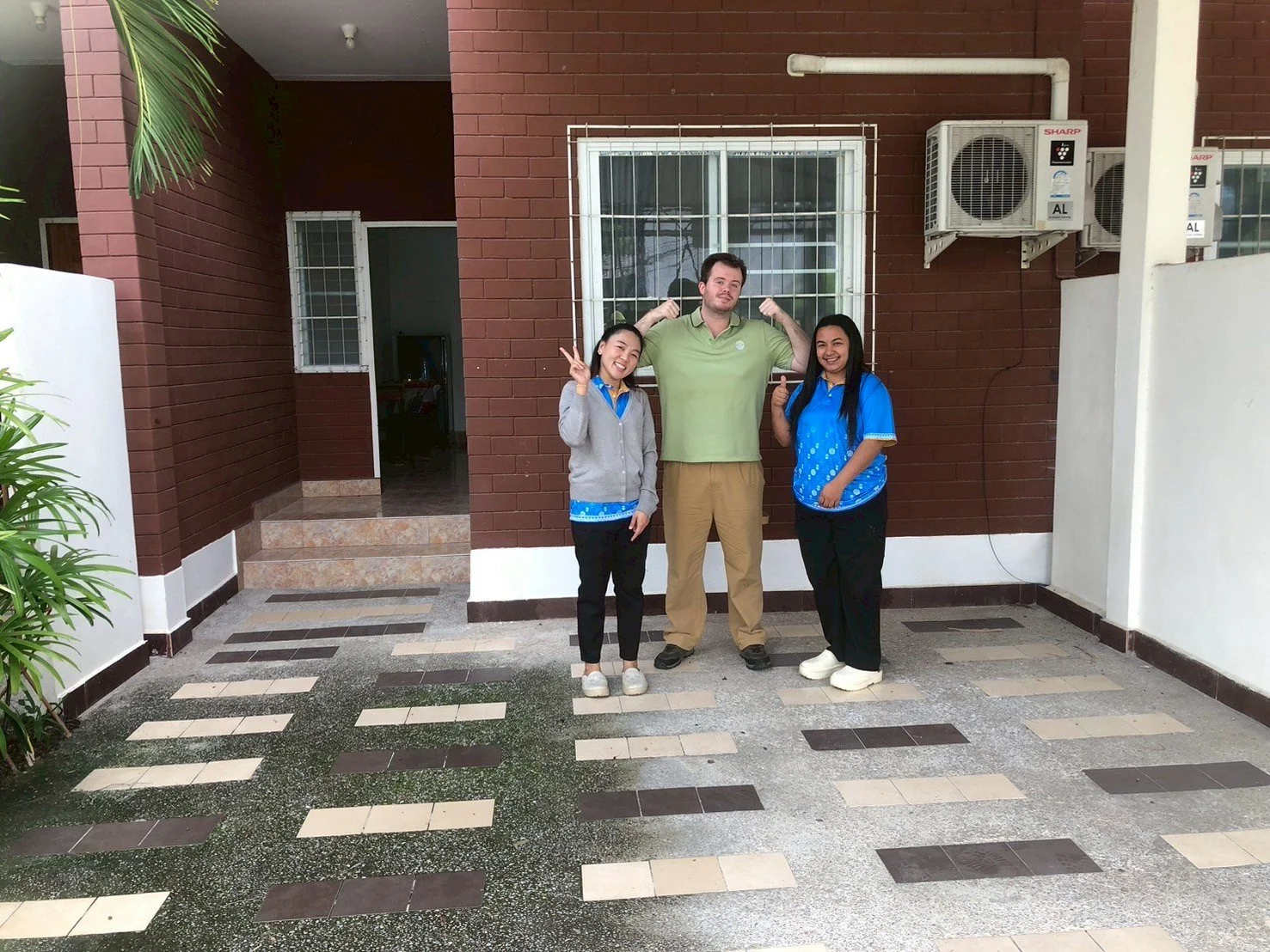 Three people from Kids English Thailand standing outside a building, one man flexing his arms, two women smiling, with air conditioning units visible on the wall behind them.
