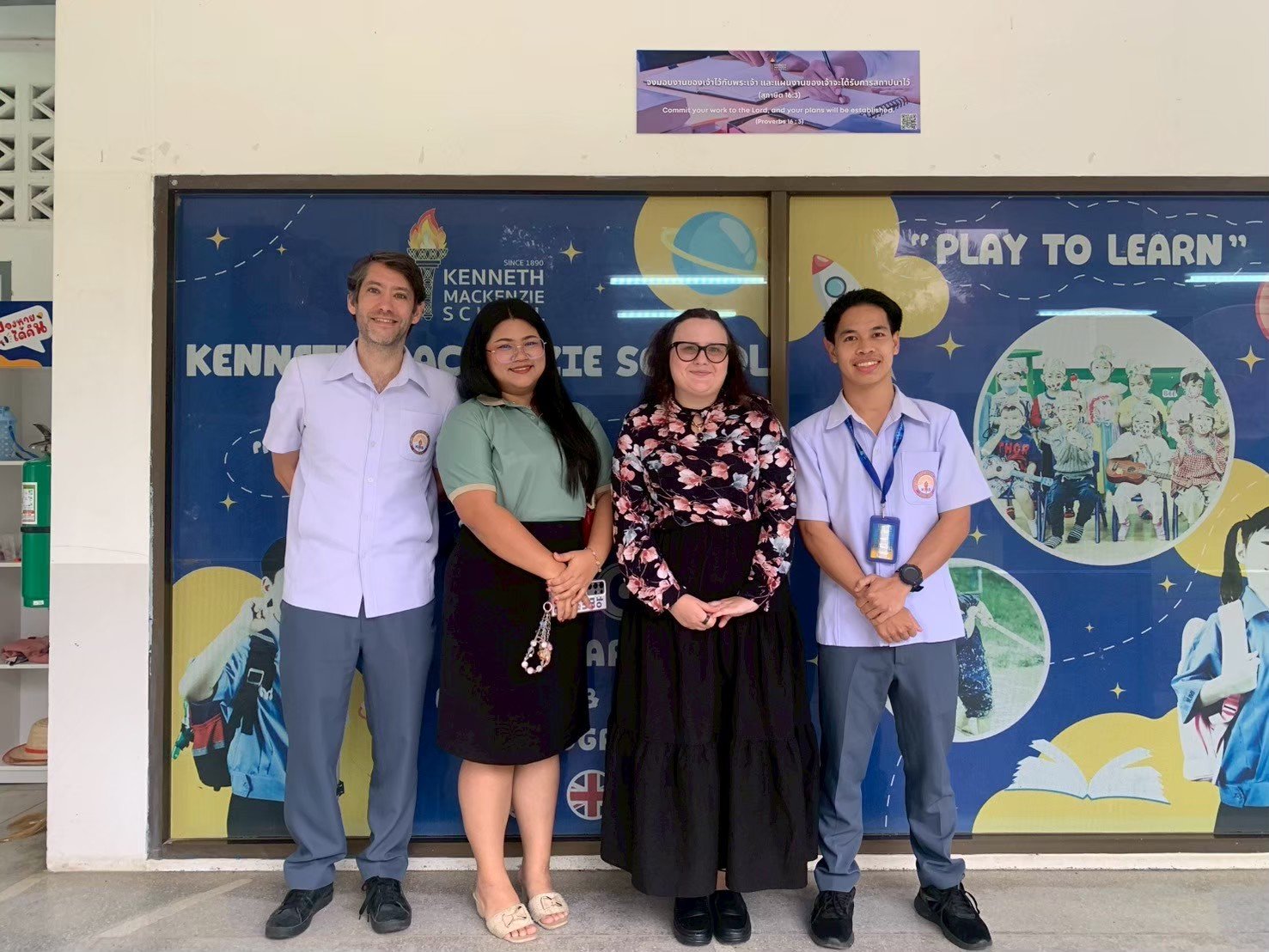 Four people from from Kids English Thailand standing in front of a school window with a colorful educational poster. 