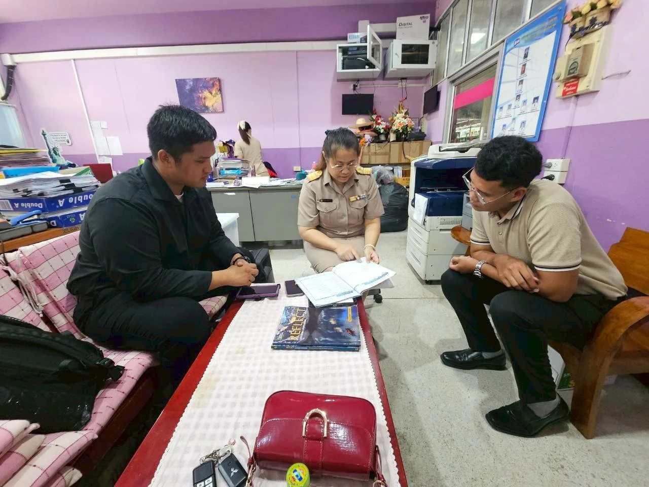 Three people from Kids English Thailand in an office engaged in a discussion, with documents open on the table. Two men are seated on either side, and a woman in uniform sits between them.