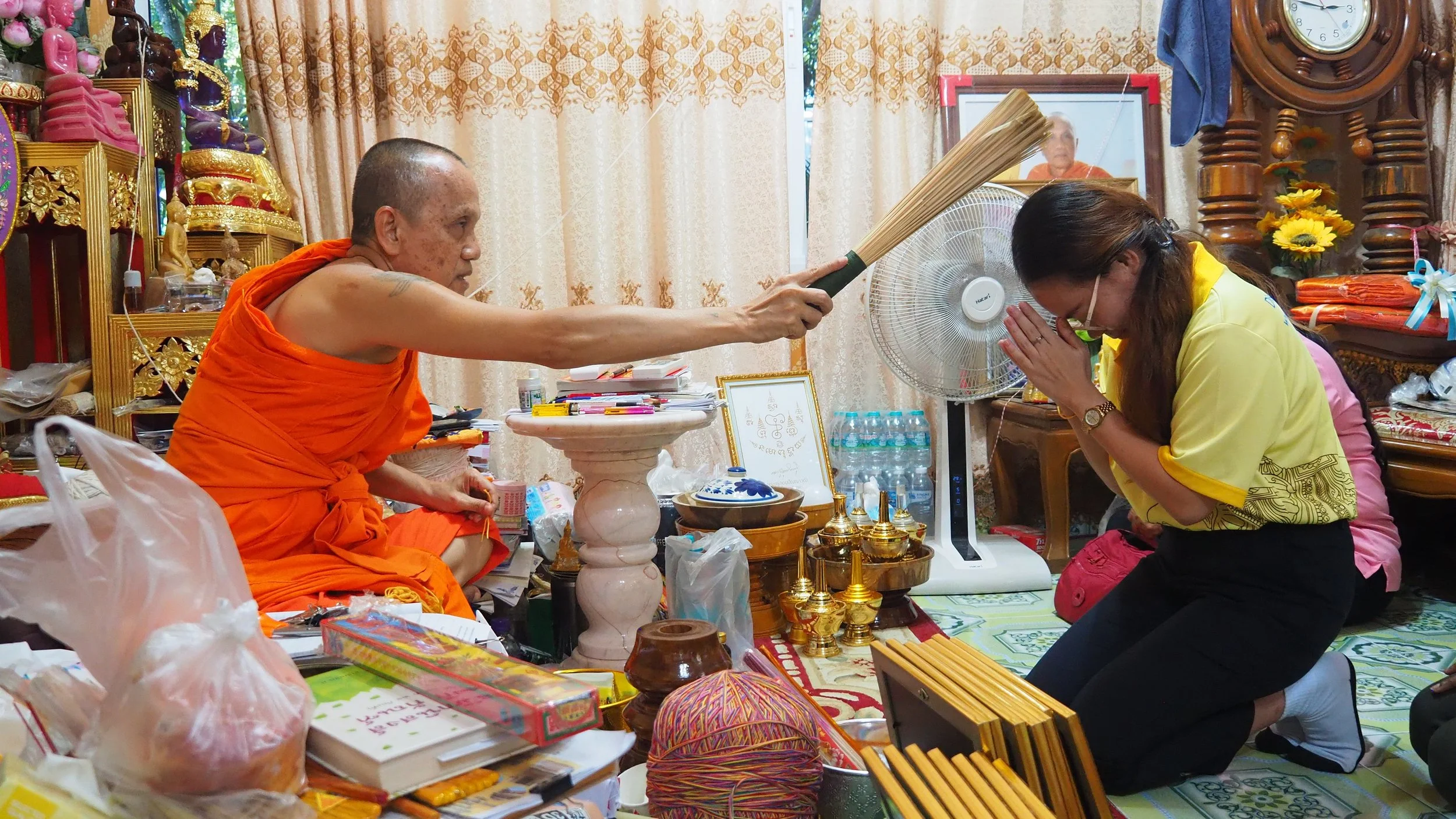 A Buddhist monk in orange robes performs a blessing or prayer ritual for a Kids English Thailand teacher in a room decorated with religious artifacts and various offerings.