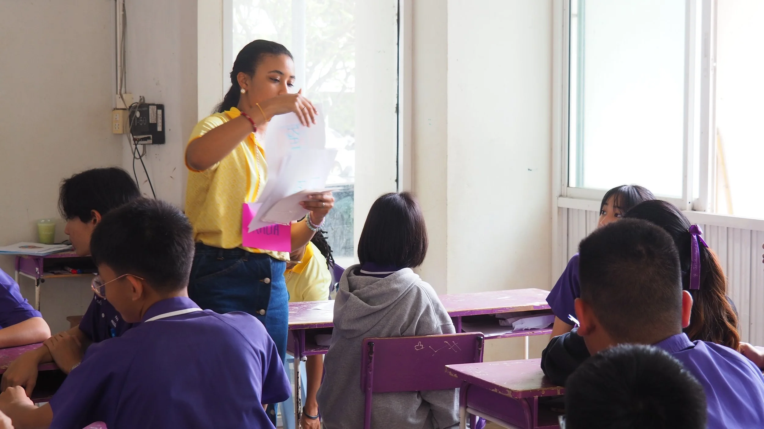 A classroom scene with a female Kids English Thailand teacher standing and speaking to students. She is holding papers and appears to be explaining something. 