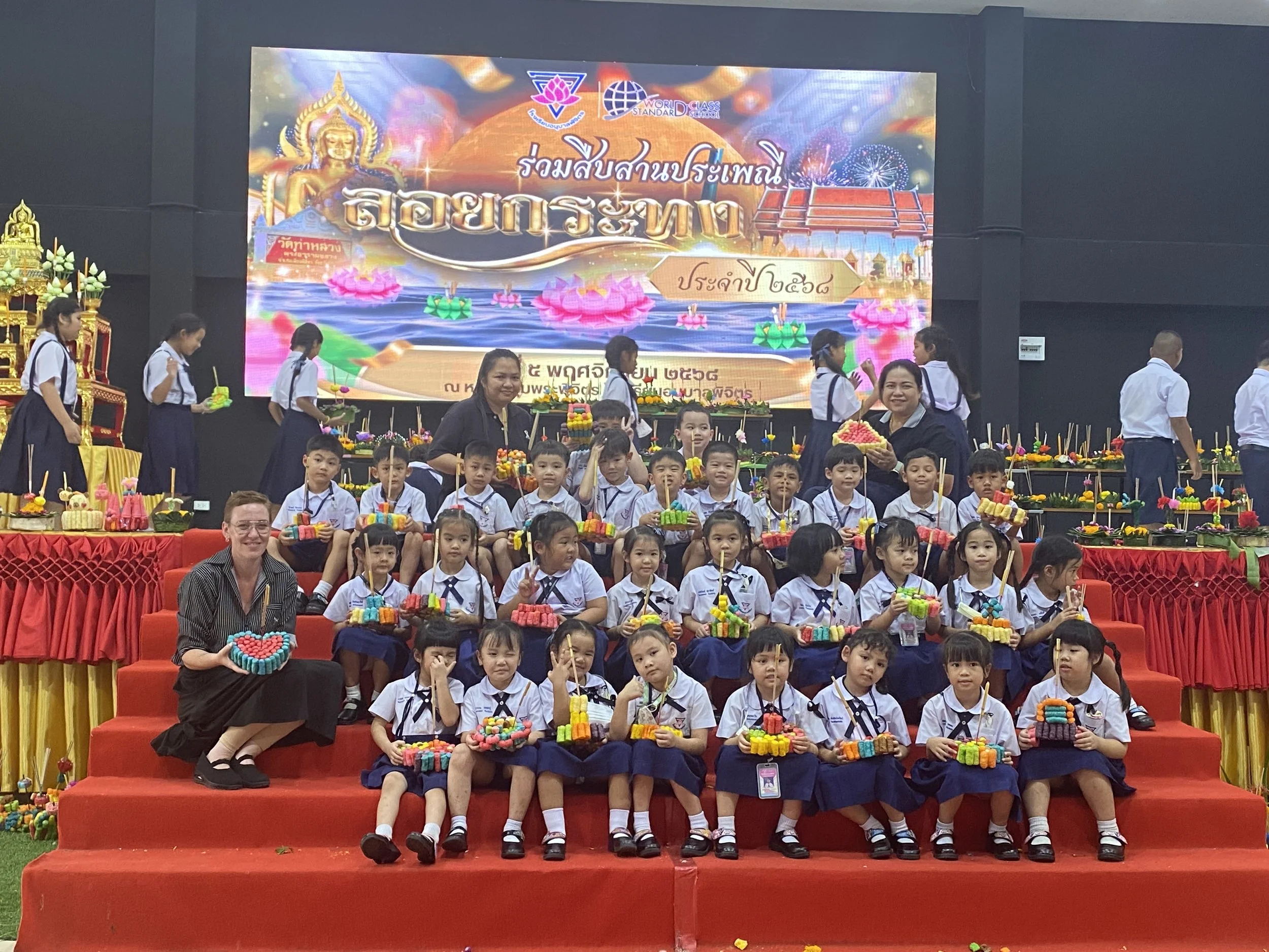 A group of young students in school uniforms sitting on red steps, holding colorful paper craft baskets, with Kids English Thailand teachers  standing behind them.