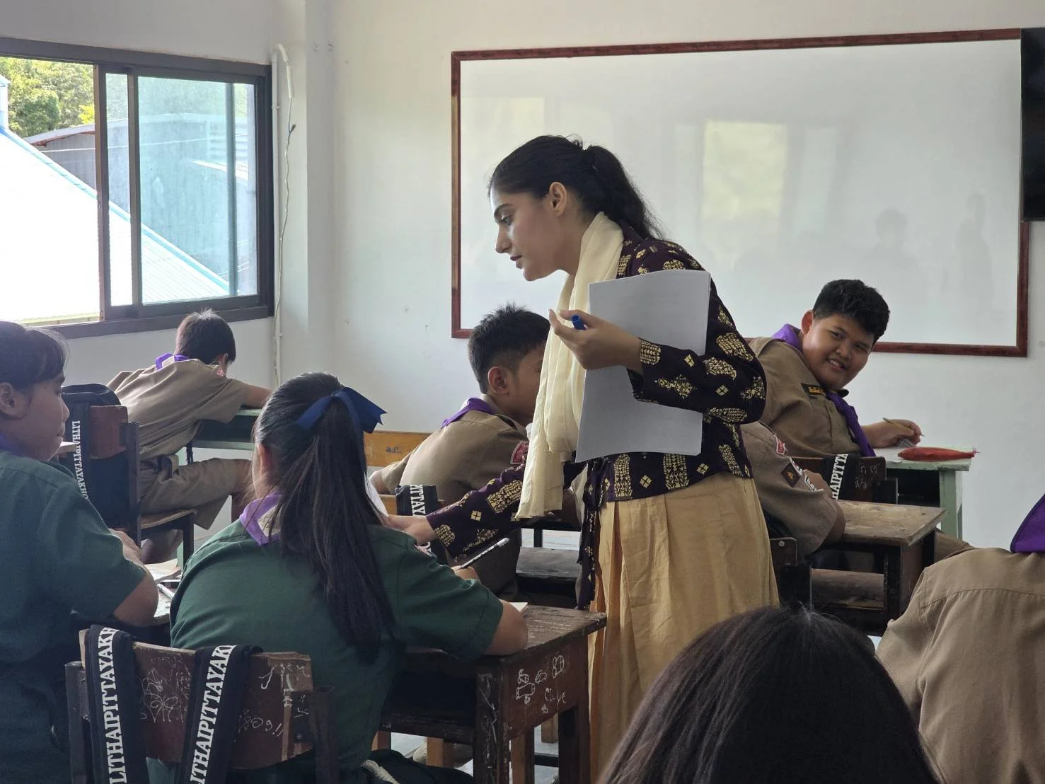 A classroom scene with a female Kids English Thailand teacher standing and speaking to students, who are seated at desks, some taking notes, inside a classroom with sunlight coming through a window.