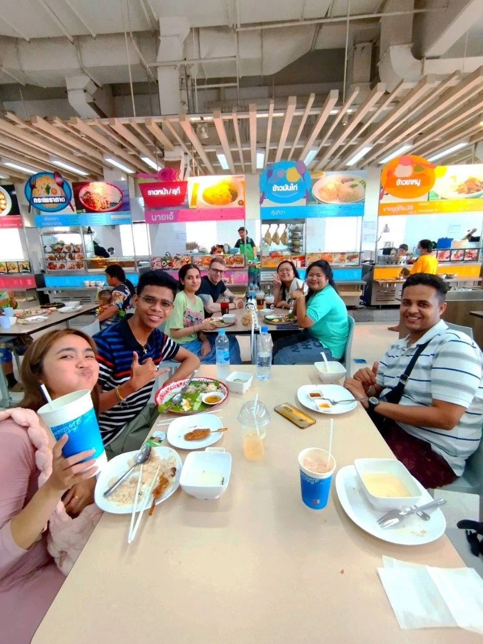 A group of eight people from Kids English Thailand dining at a food court with various food stations in the background, some holding drinks and smiling at the camera.