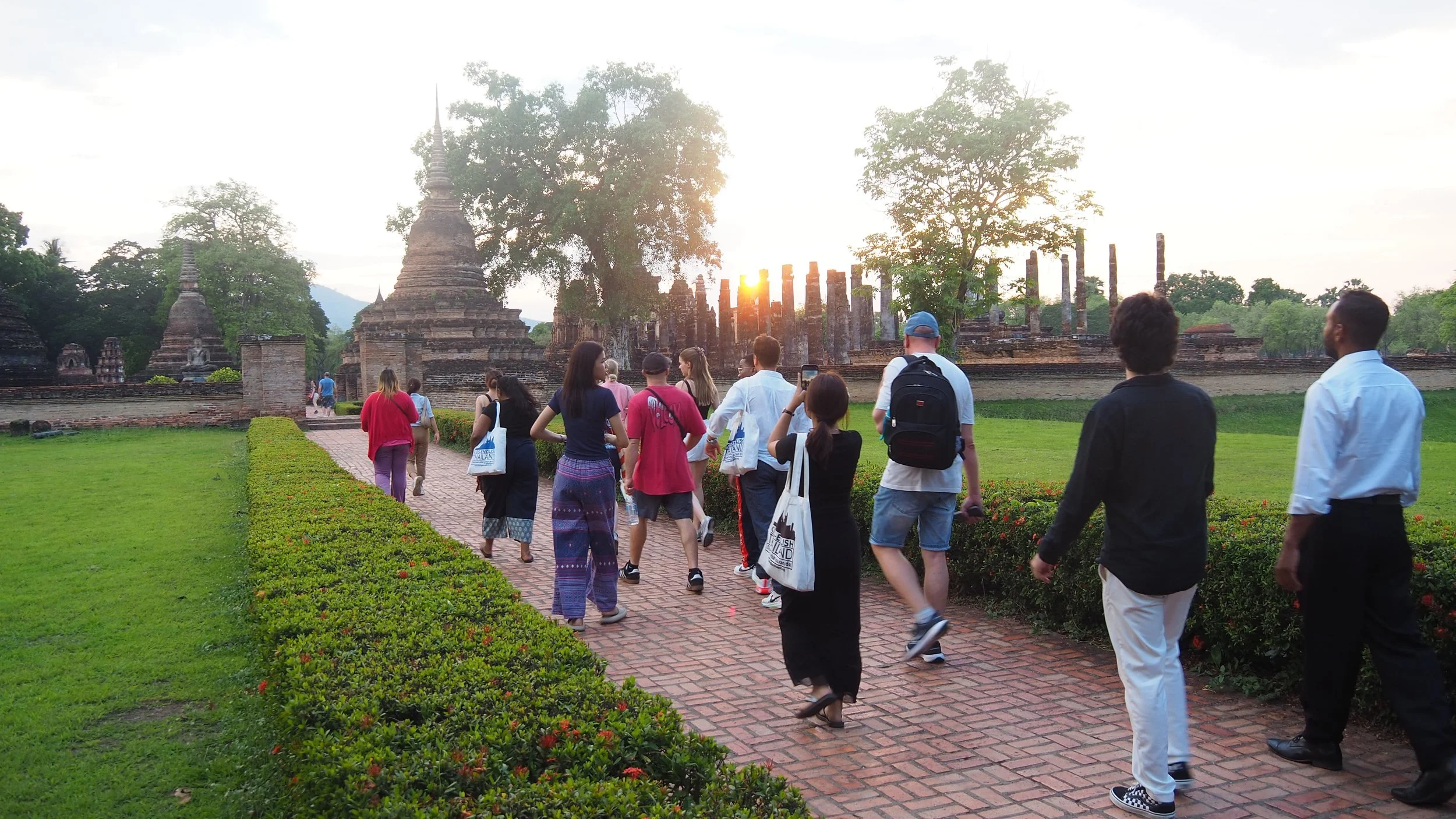 Group of Kids English Thailand teachers walking along a brick pathway towards ancient temple ruins at sunset in a lush, green park.